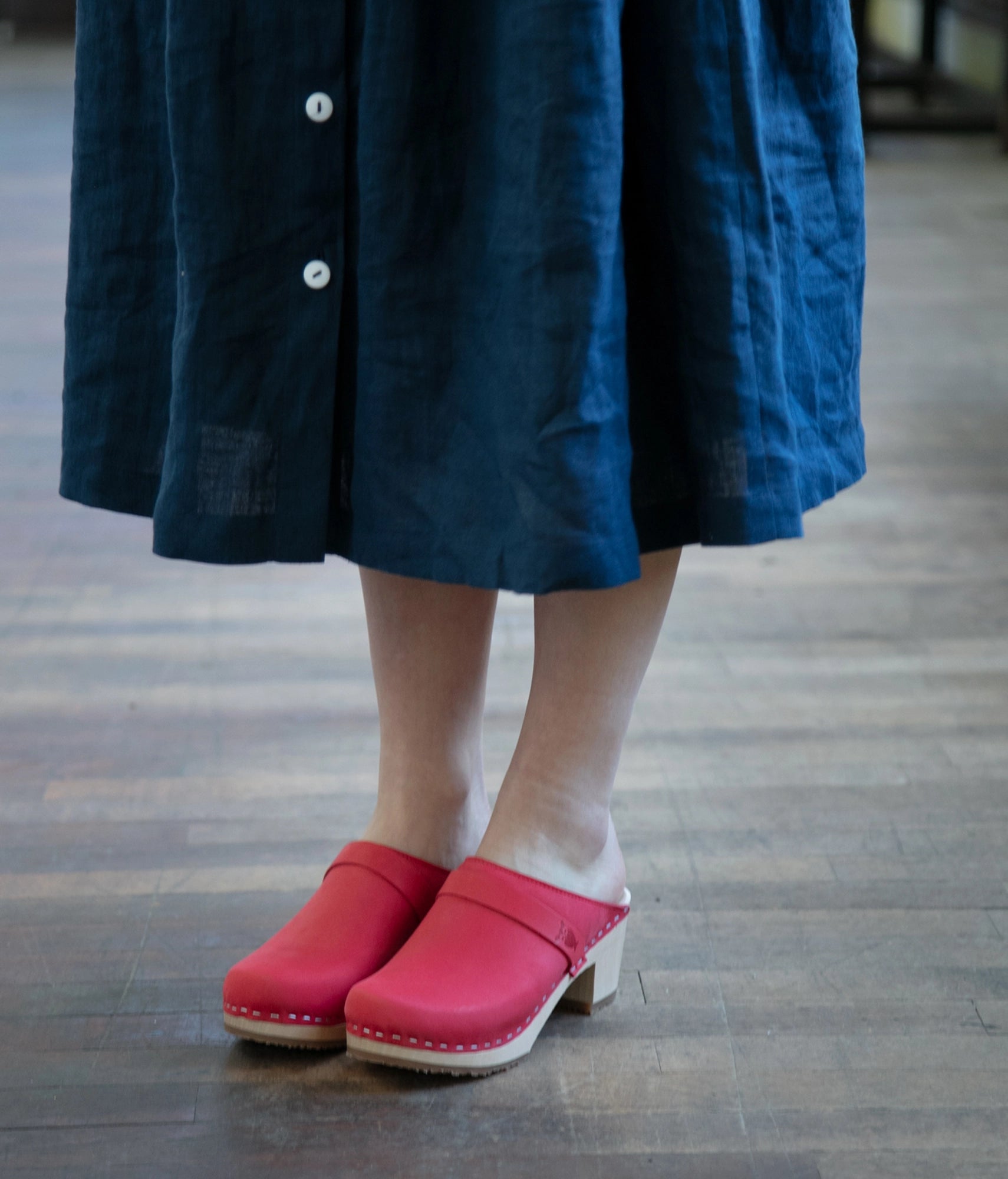 Person wearing a blue dress and red clogs on a wooden floor. classic high heeled clog mule in red nubuck leather stapled on a light wooden base with a leather strap
