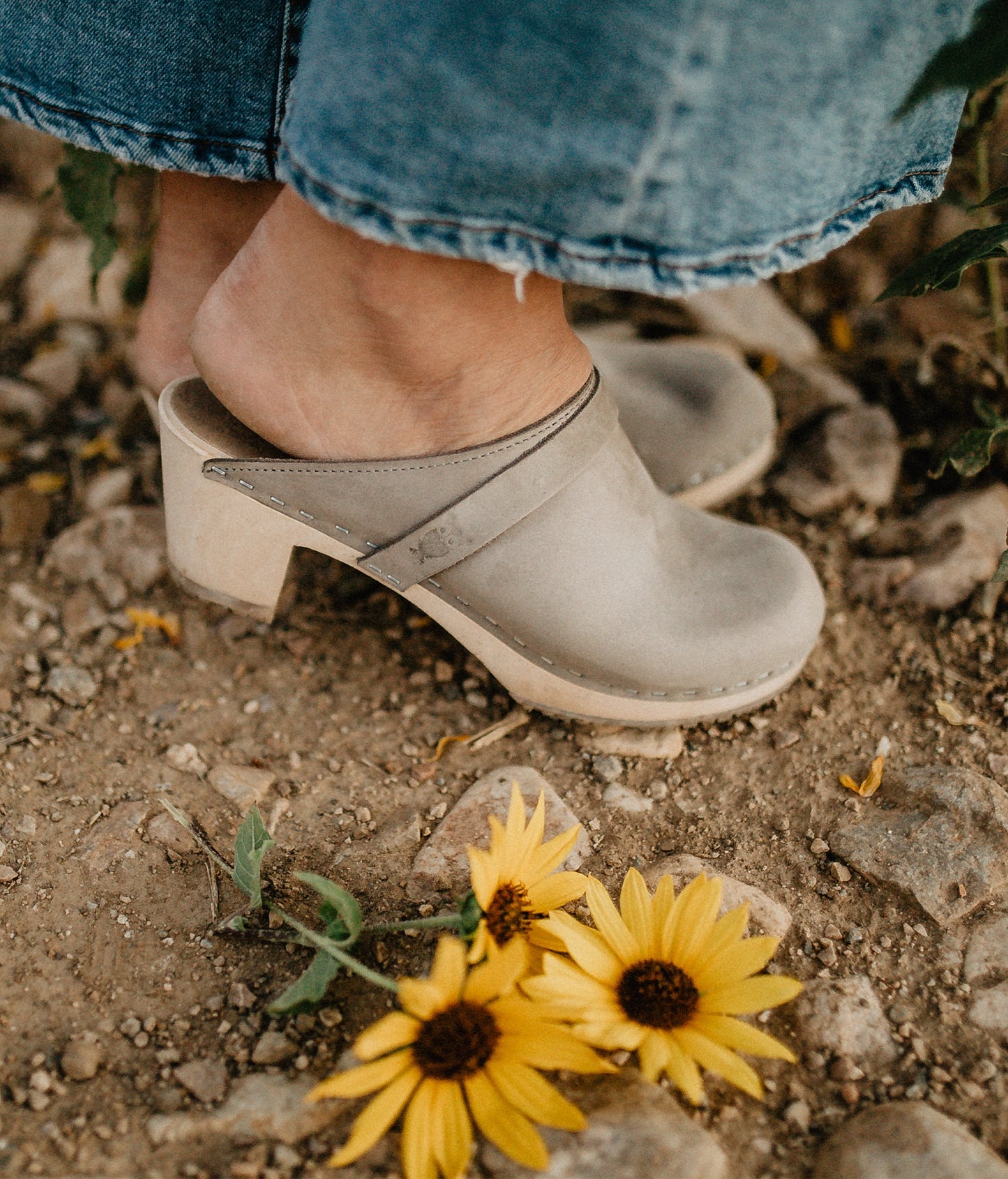 Stone colored clog shoe worn on a foot, standing on a rocky ground with yellow flowers. Classic high heeled clog mule in stone grey nubuck leather stapled on a light wooden base with a leather strap