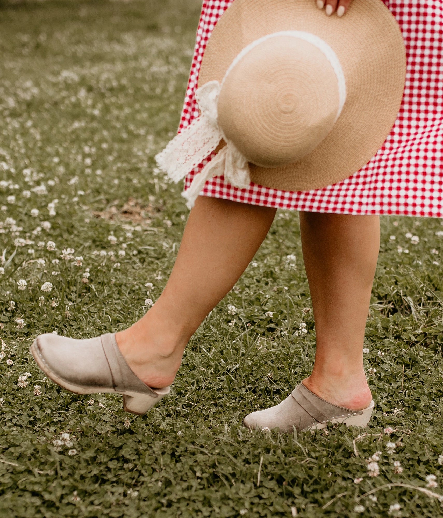 Person in a checkered dress walking on grass with flowers. Classic high heeled clog mule in stone grey nubuck leather stapled on a light wooden base with a leather strap