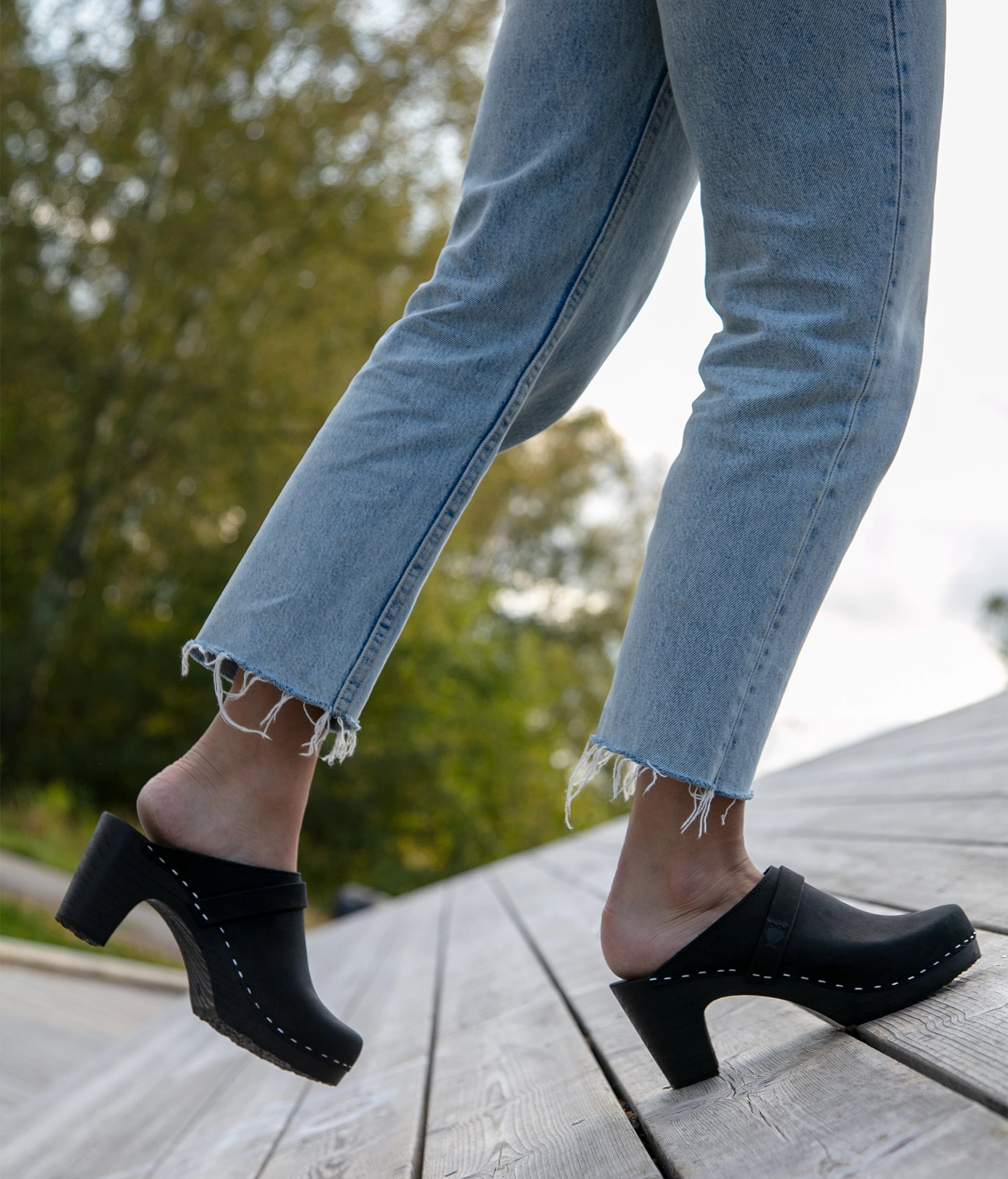 Person wearing all black clogs and light blue jeans on a wooden deck with blurred greenery in the background