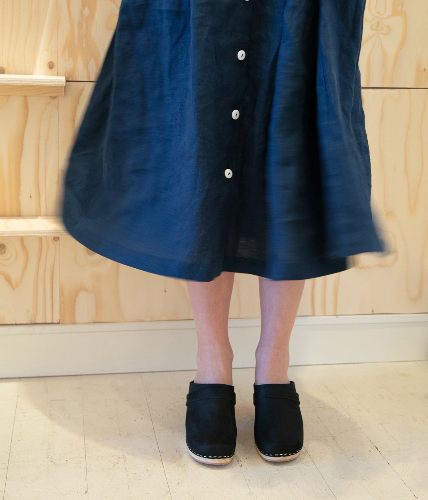 Person wearing a navy button-up dress with black clogs against a wooden background