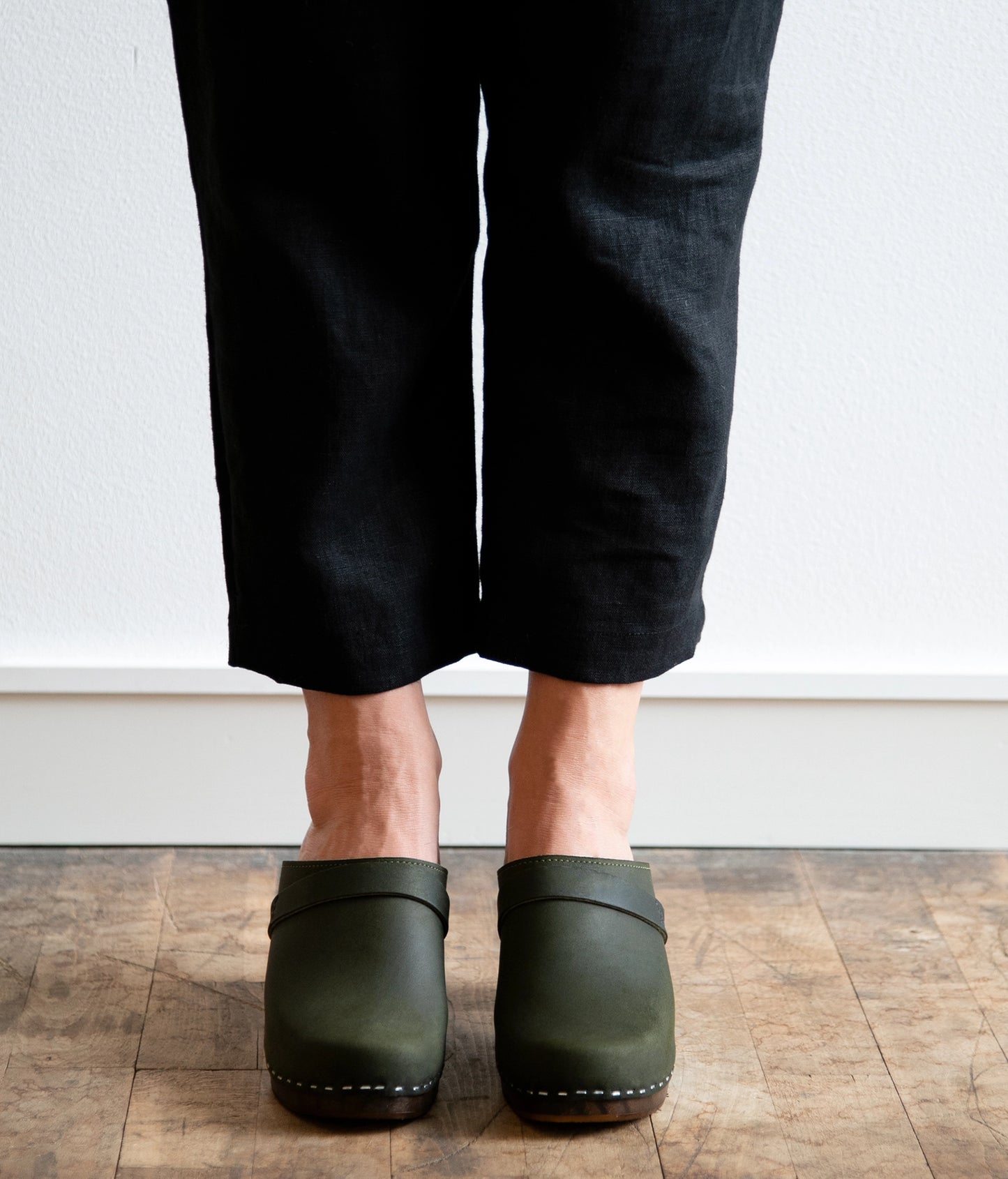 Person wearing black pants and Olive green clog mules on a wooden floor.