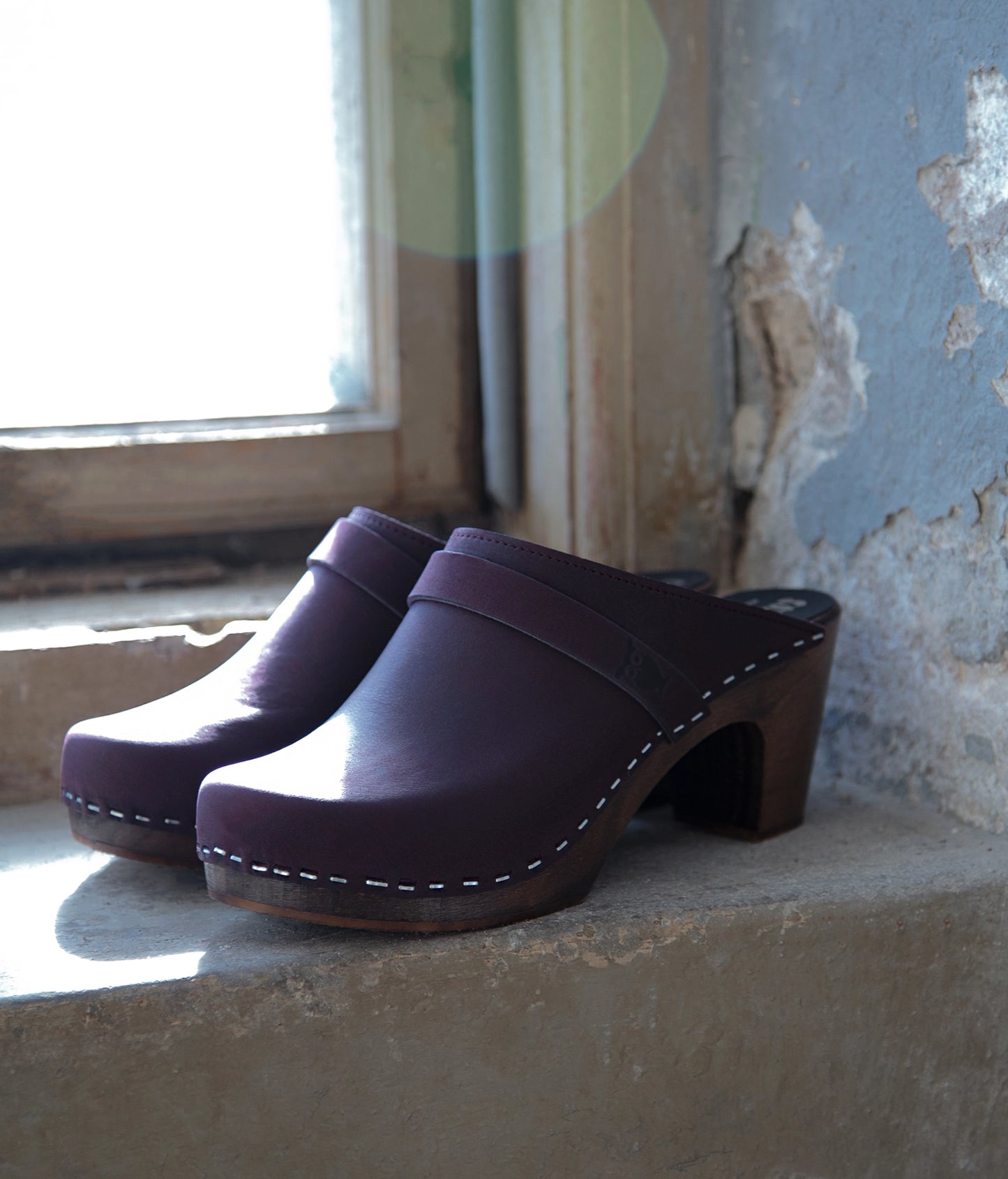Pair of plums colored clog mules on a windowsill with peeling paint.