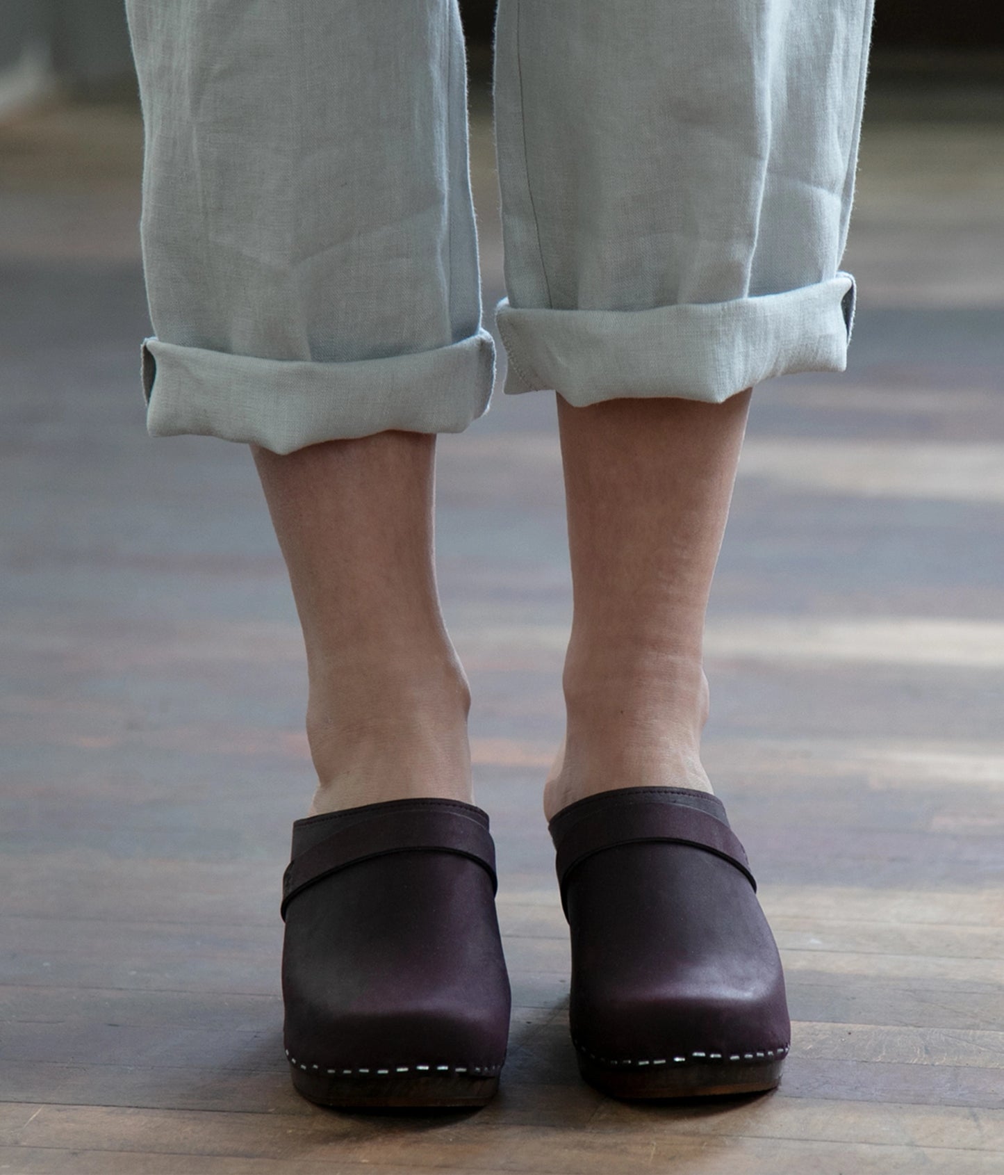 Plum colored leather clogs worn with light-colored pants on a wooden floor.
