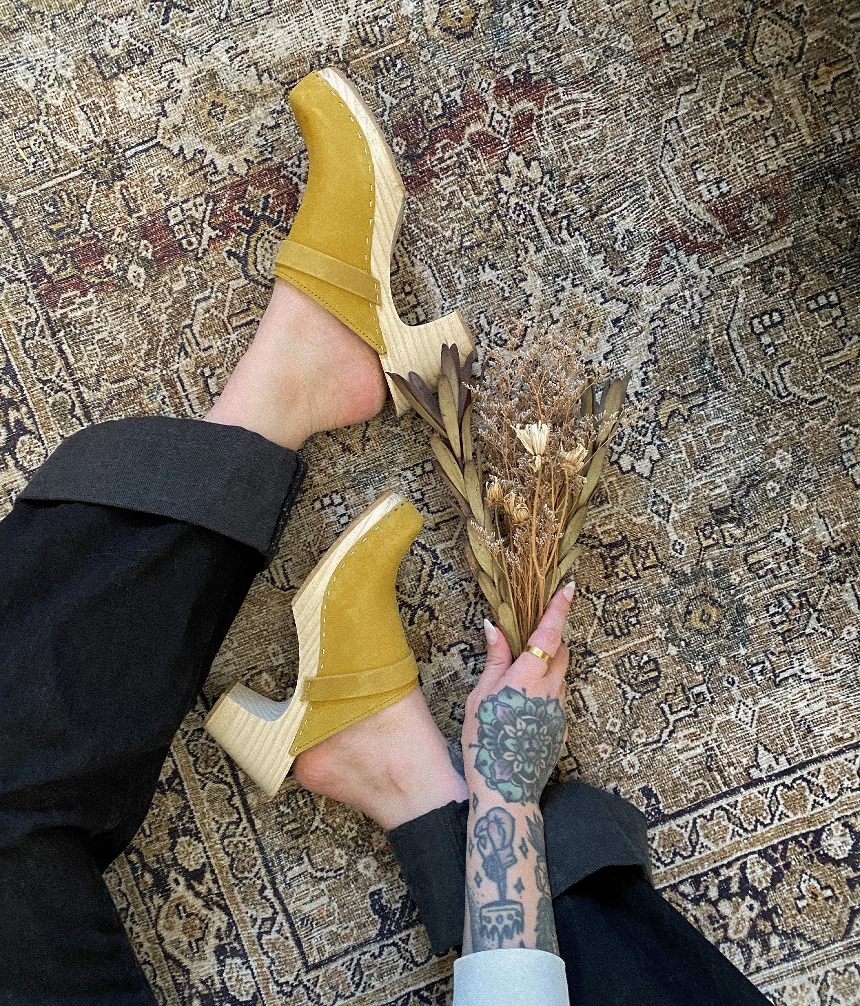 Person wearing Royal yellow clog mules holding a dried flower bouquet on a patterned rug.