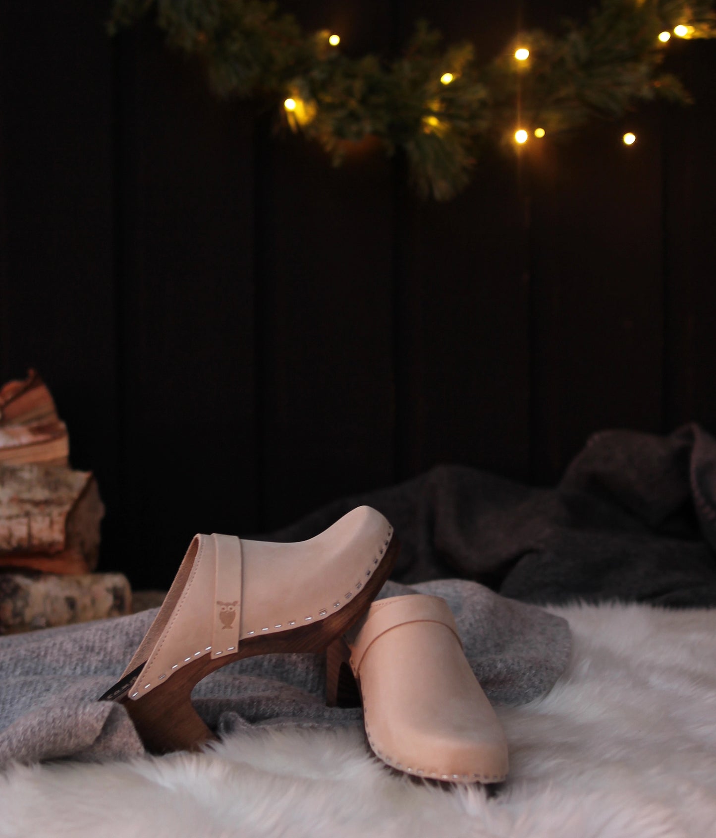 Pair of sand colored clog mules on dark base laying on a textured surface with a dark background and string lights.