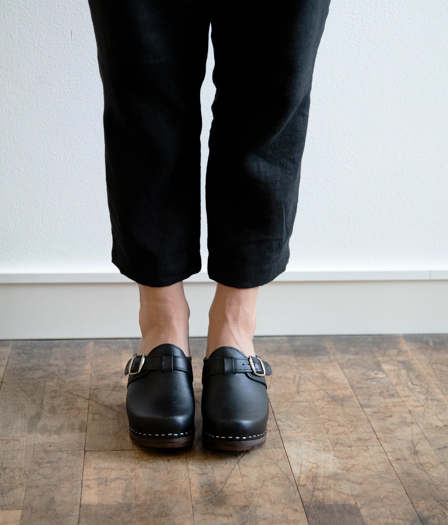 high heeled clog mules in black vegetable tanned leather with a silver buckle stapled on a dark wooden base