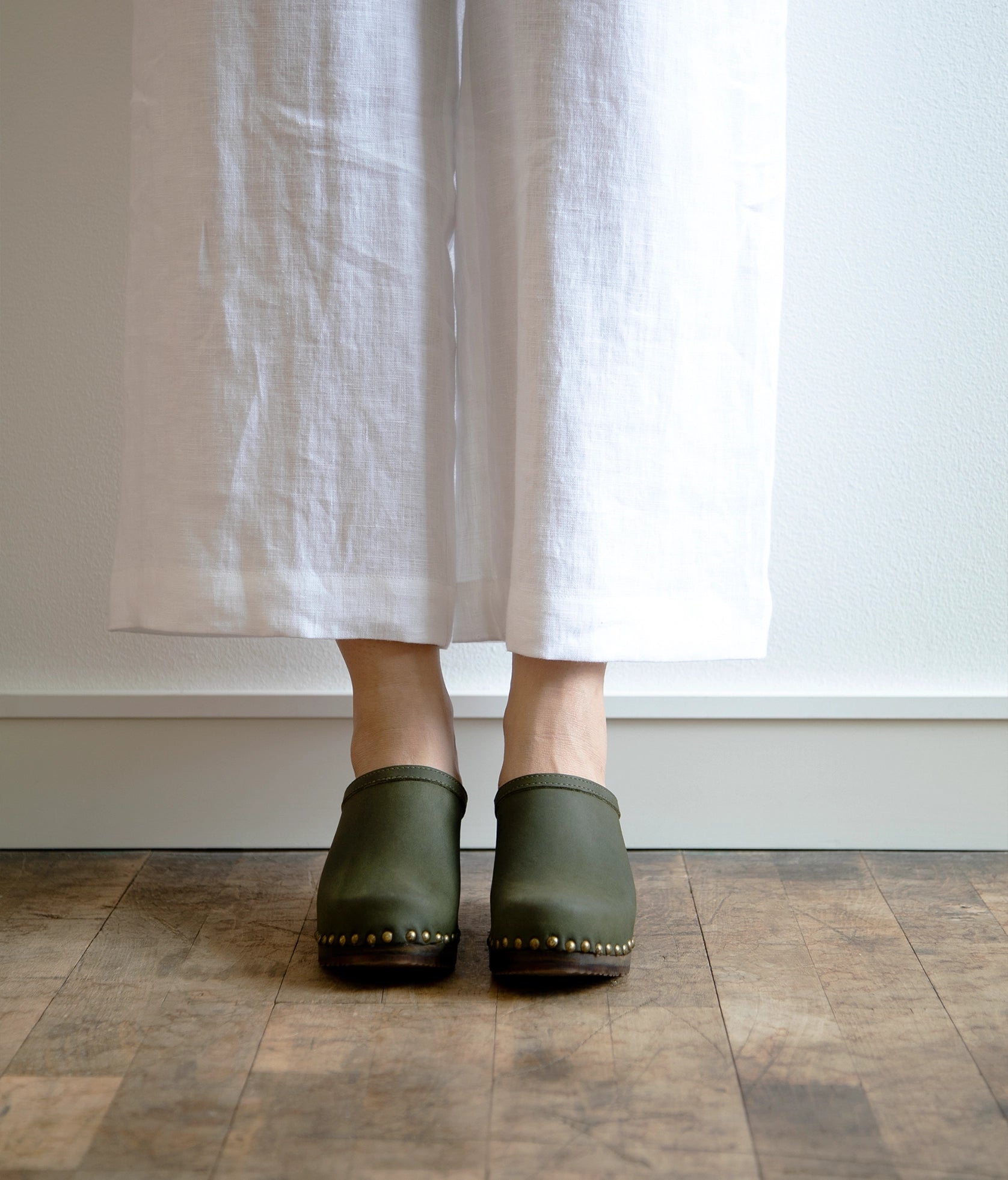 high heeled clog mules in olive green nubuck leather stapled on a dark wooden base with brass gold studs