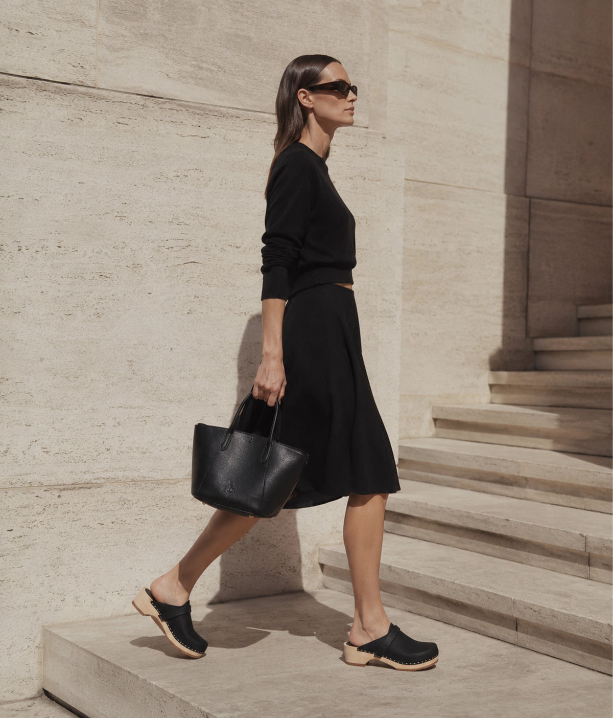 Woman in black outfit with a black bag walking on stone steps with black clogs