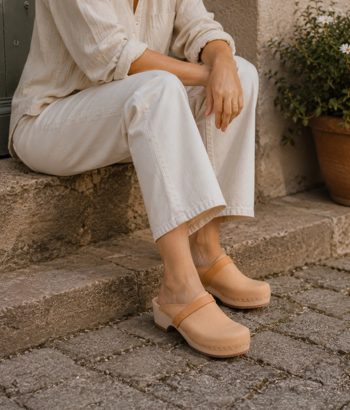 Person wearing beige clogs sitting on stone steps with a plant in the background