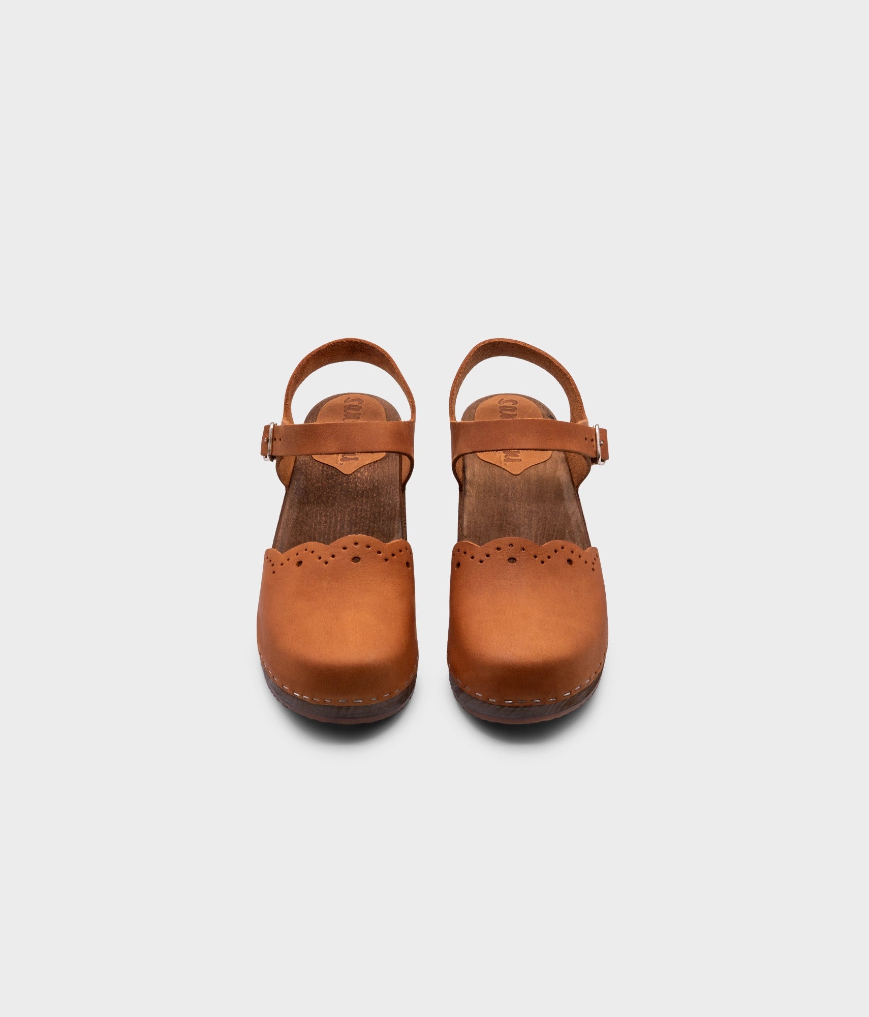 low heeled closed-toe clog sandals in light brown nubuck leather with a wavy leather cutout stapled on a dark wooden base