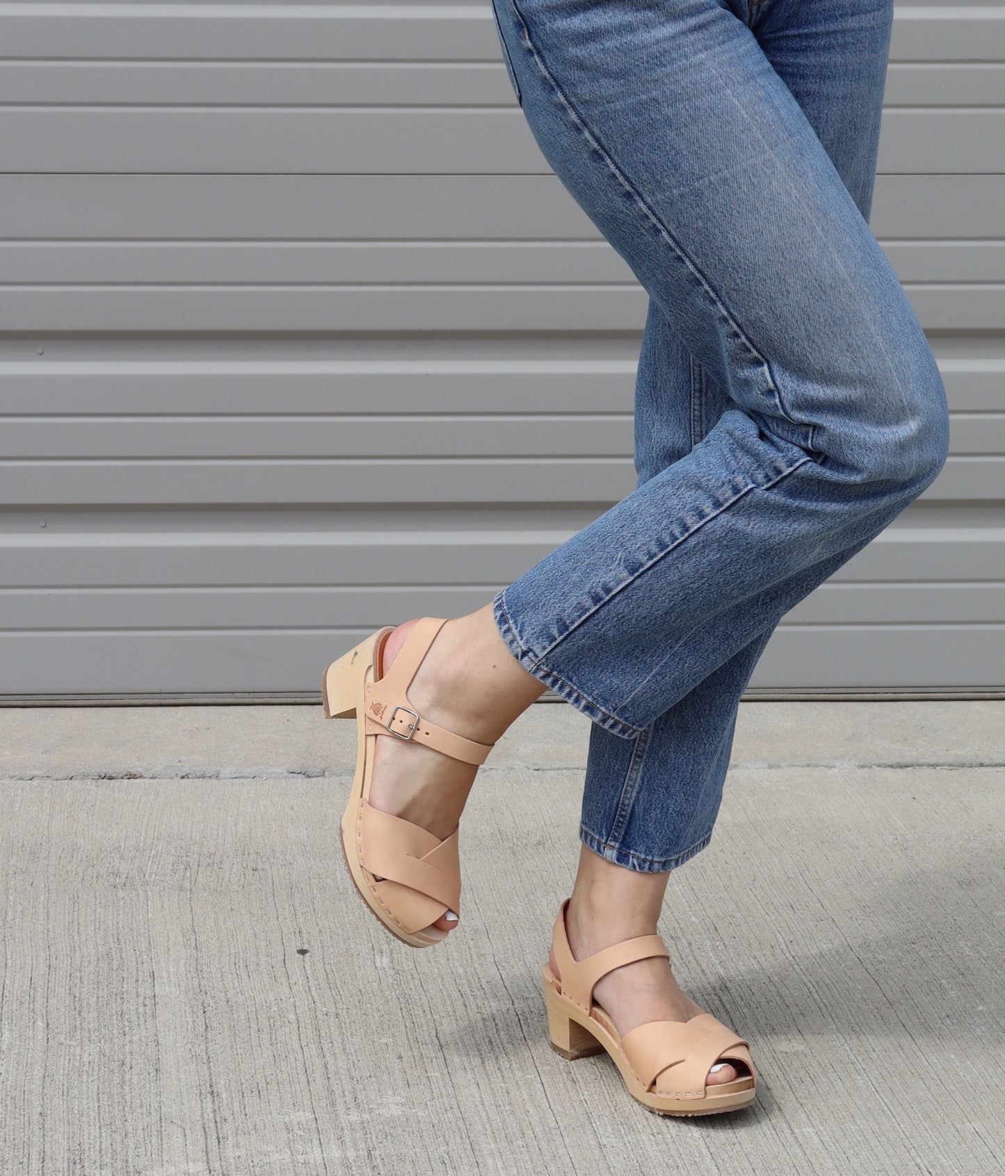 Woman  wearing high heeled clog sandals in ecru beige vegetable tanned leather with an open-toe stapled on a light wooden base