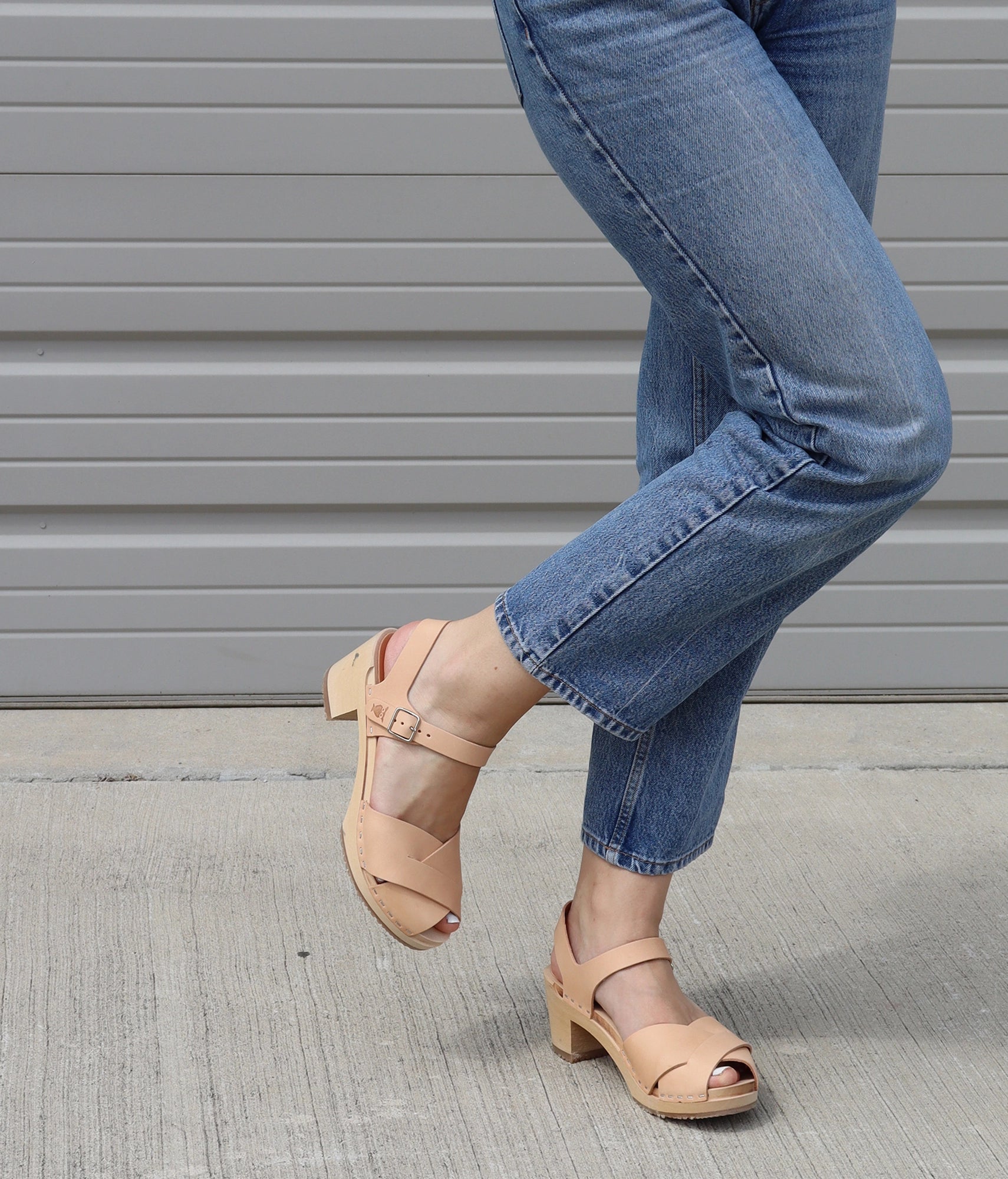 Woman  wearing high heeled clog sandals in ecru beige vegetable tanned leather with an open-toe stapled on a light wooden base