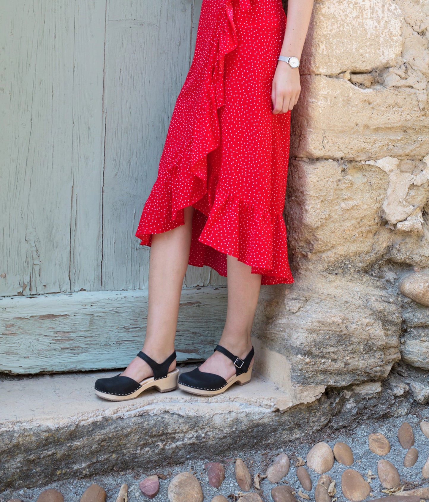 woman wearing classic low heeled closed-toe sandal in black nubuck leather stapled on a light wooden base with a red skirt