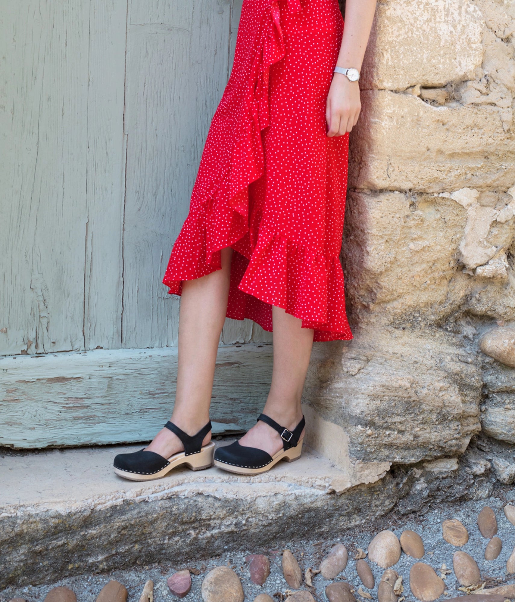 woman wearing classic low heeled closed-toe sandal in black nubuck leather stapled on a light wooden base with a red skirt