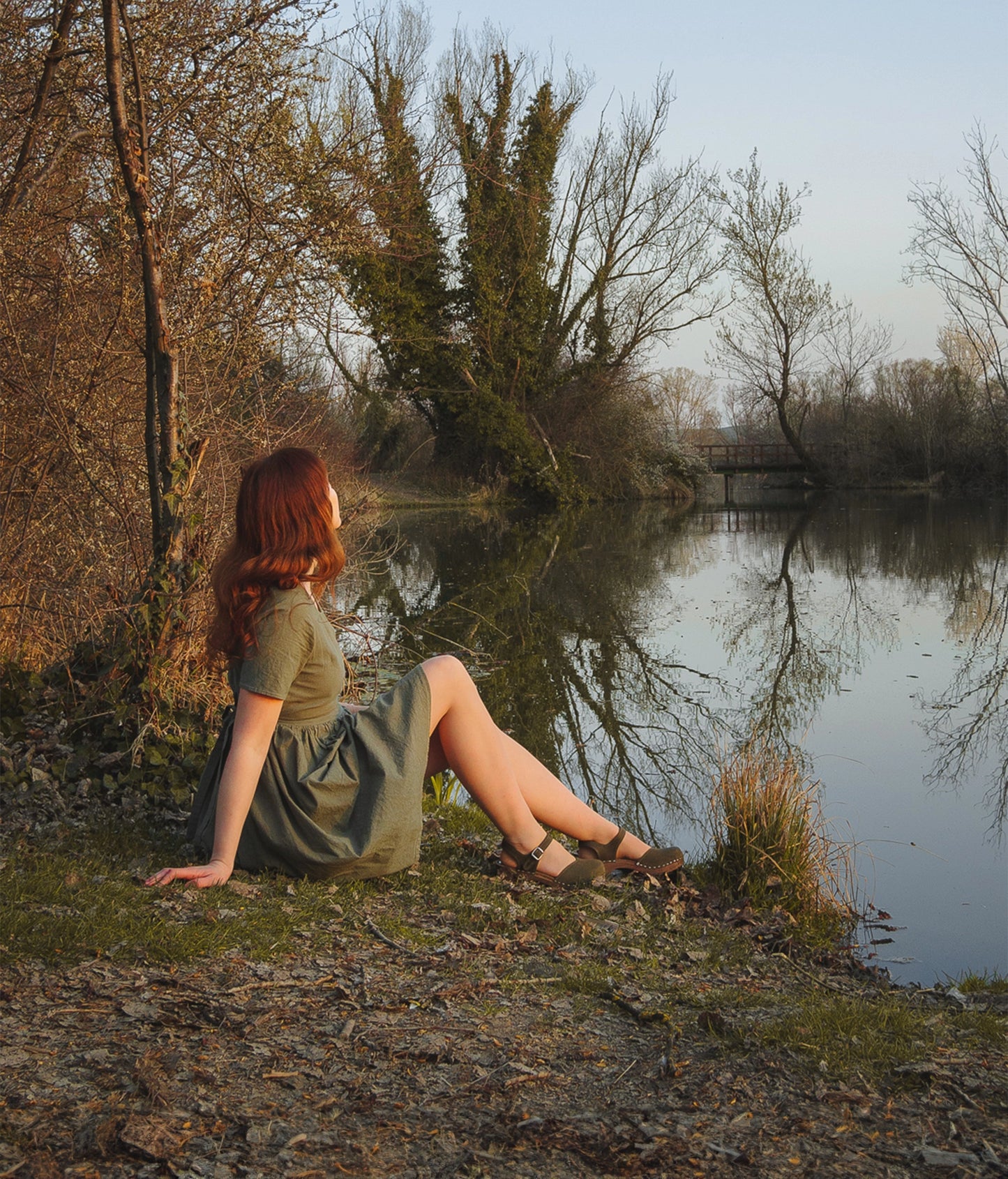 woman wearing a green midi dress and classic low heeled closed-toe sandal in olive green nubuck leather stapled on a dark wooden base