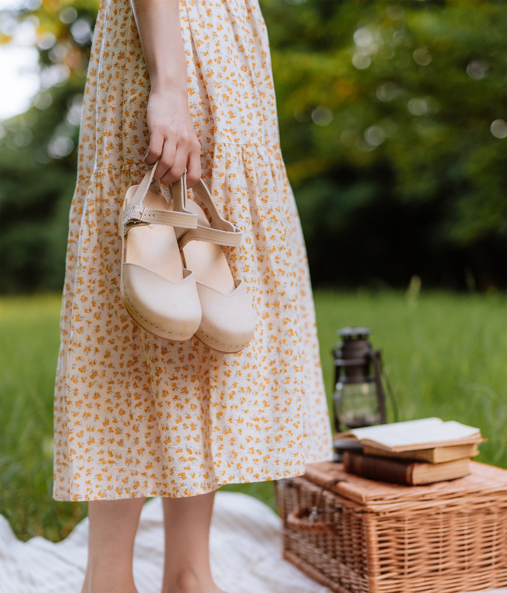 woman holding a pair of classic low heeled closed-toe sandal in sand white nubuck leather stapled on a light wooden base