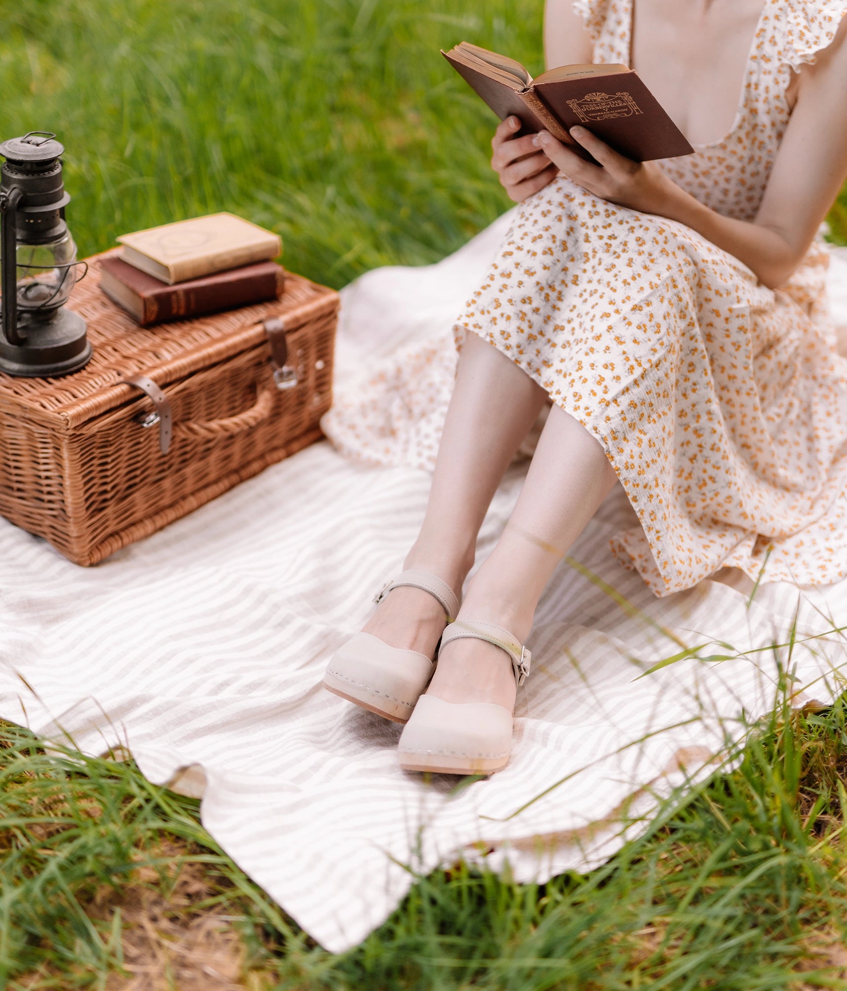 woman wearing a pair of classic low heeled closed-toe sandal in sand white nubuck leather stapled on a light wooden base with a white flowery dress