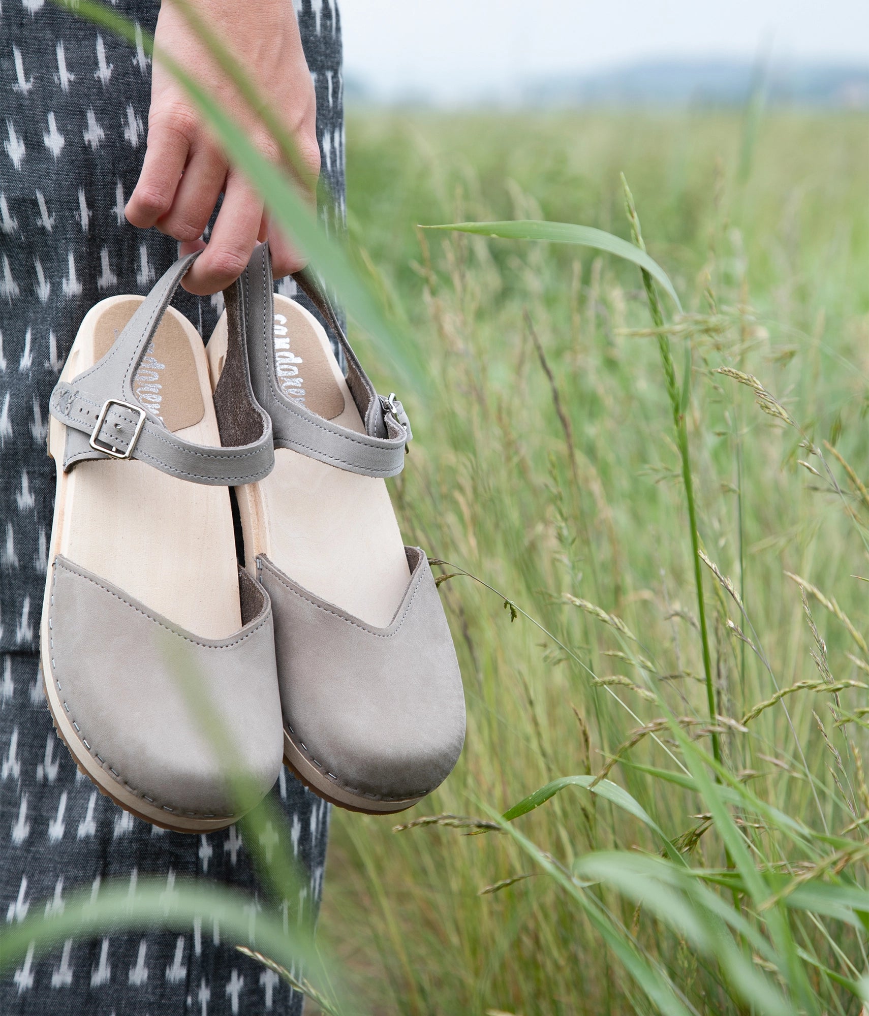 Woman holding up a pair of classic low heeled closed-toe sandal in stone grey nubuck leather stapled on a light wooden base
