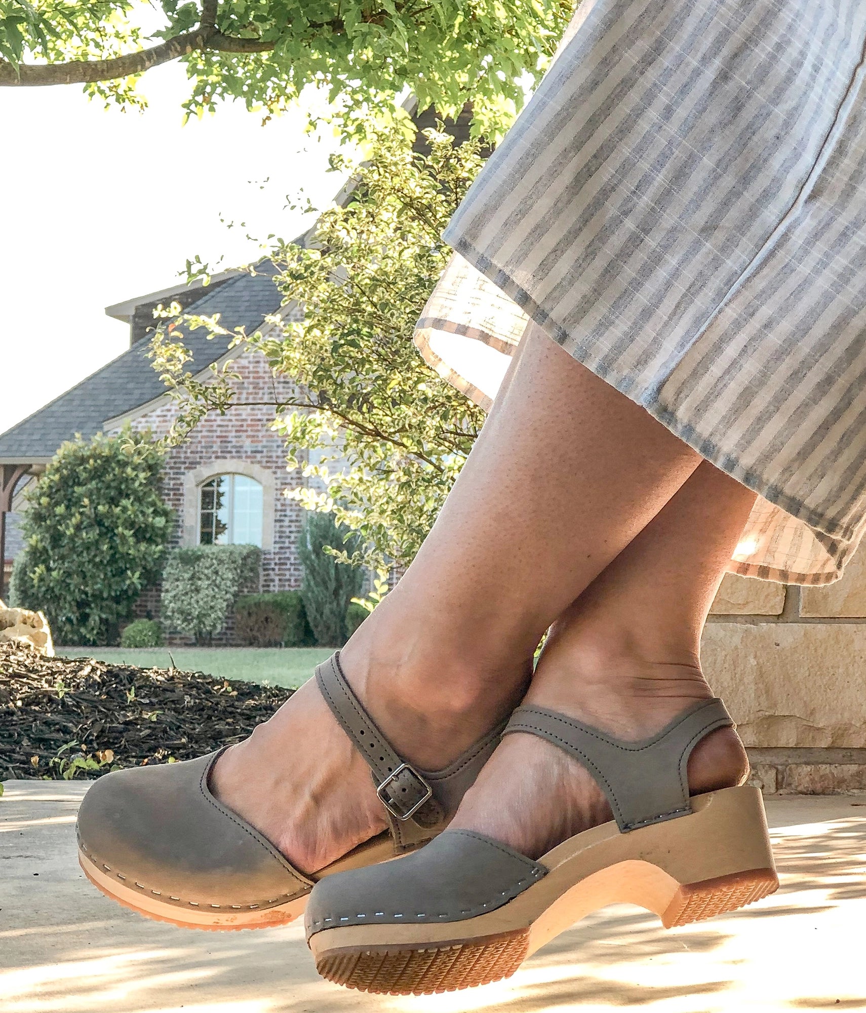 Woman wearingclassic low heeled closed-toe sandal in stone grey nubuck leather stapled on a light wooden base