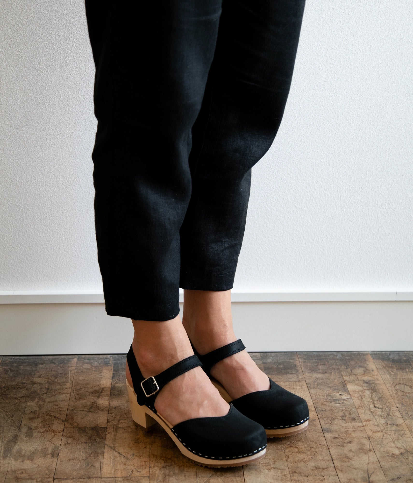 Woman wearing high heeled closed-toe clog sandal in black nubuck leather stapled on a light wooden base