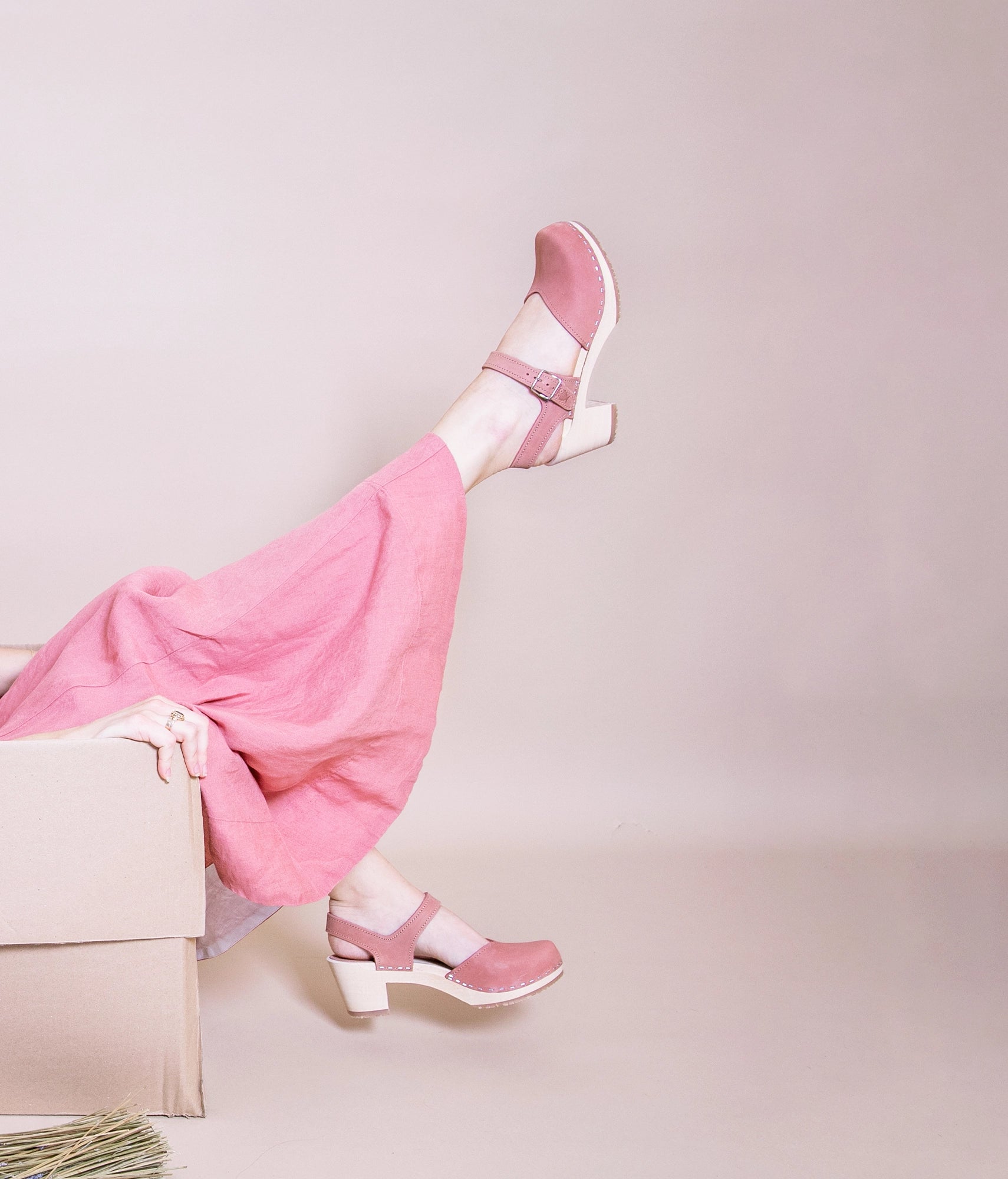 woman wearing a pink dress and a pair of high heeled closed-toe clog sandal in blush pink nubuck leather stapled on a light wooden base