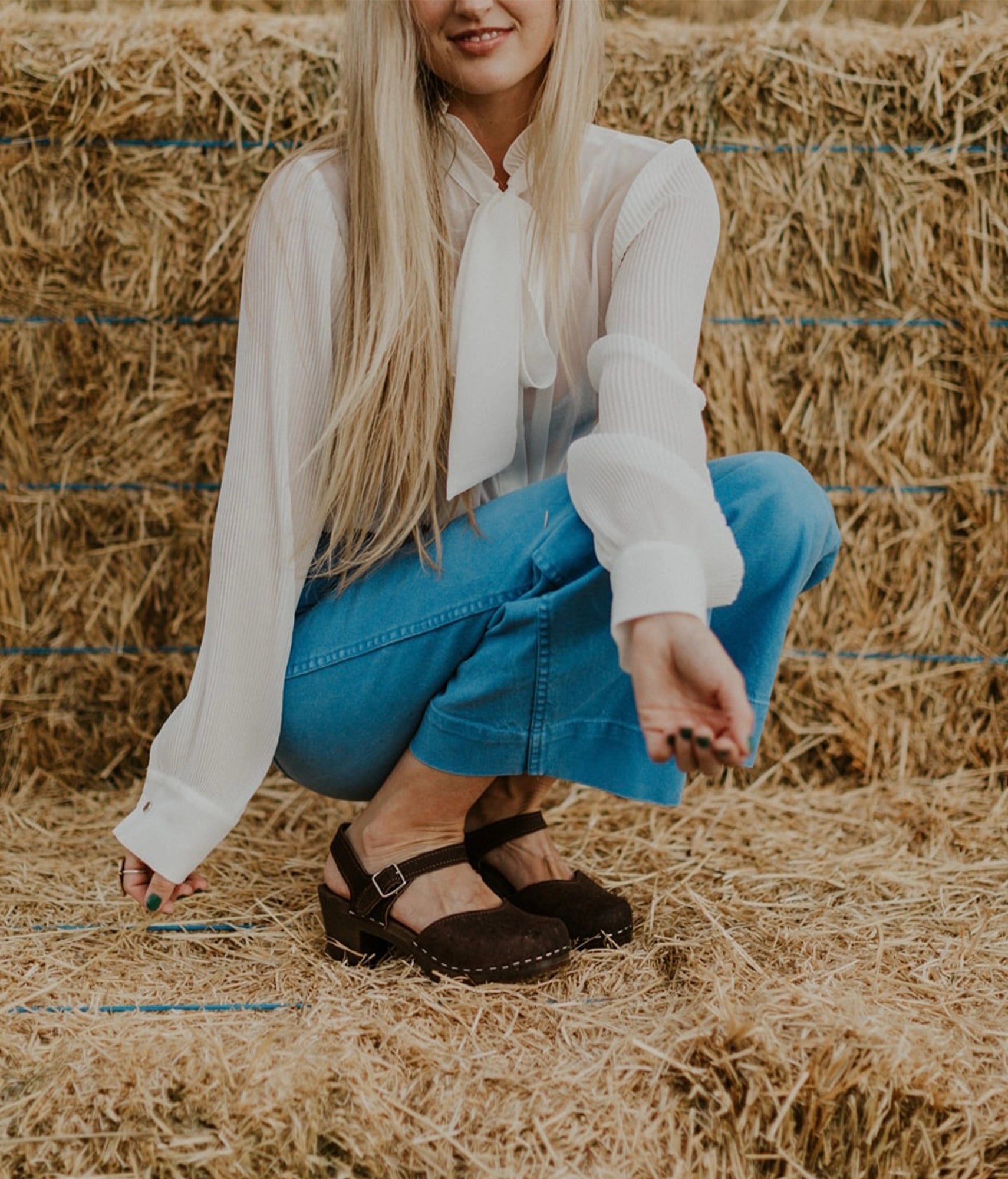 woman wearing a white blouse, blue jeans and high heeled closed-toe clog sandal in dark brown nubuck leather stapled on a dark wooden base