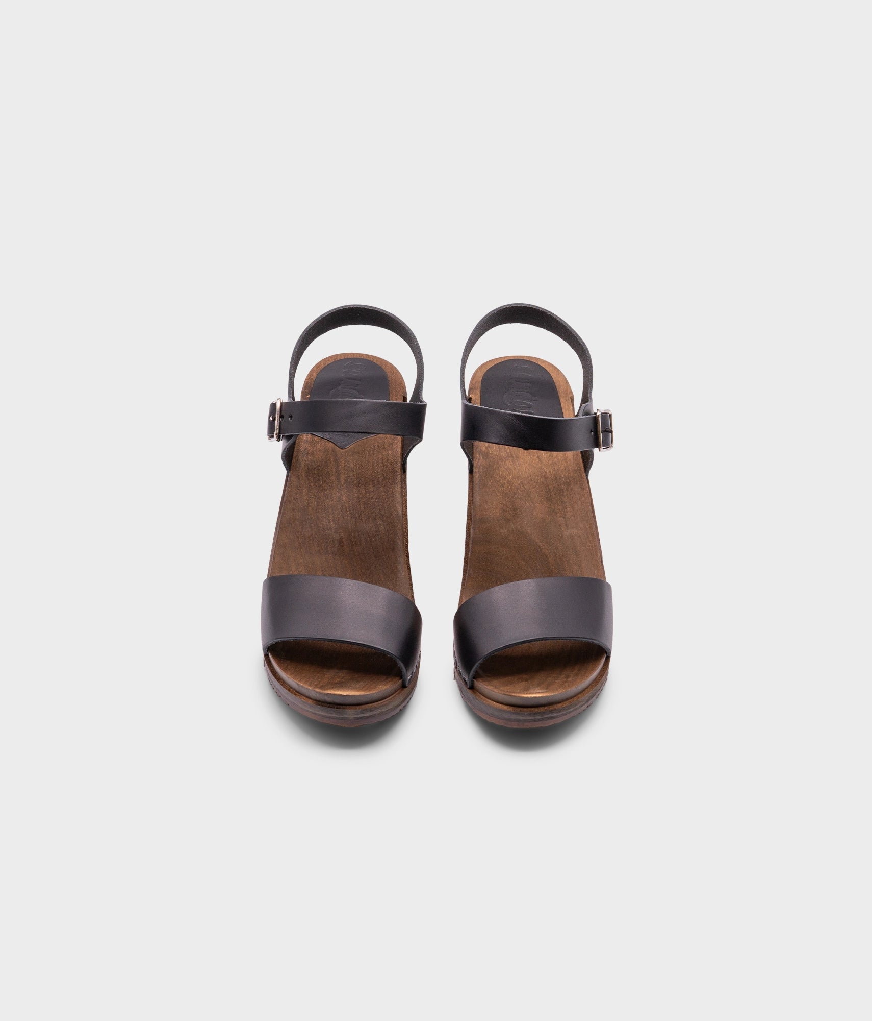 high-heeled open-toe clog sandal in black vegetable tanned leather stapled on a dark wooden base