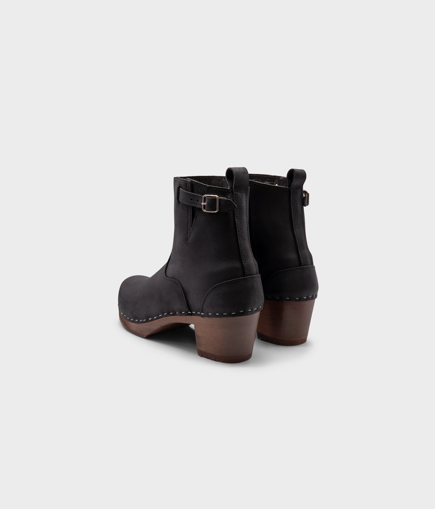 high-heeled clog boots in black nubuck leather stapled on a dark wooden base with a silver buckle
