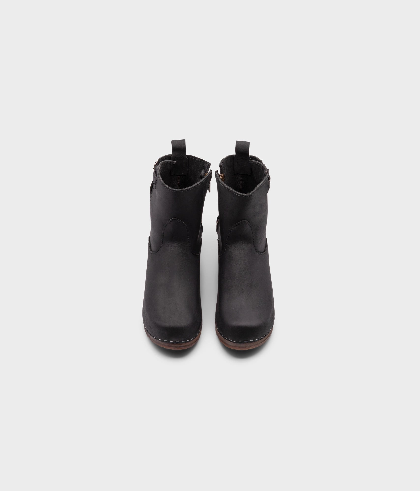 high-heeled clog boots in black nubuck leather stapled on a dark wooden base with a silver buckle