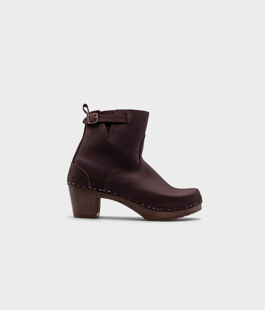 high-heeled clog boots in dark brown nubuck leather stapled on a dark wooden base with a silver buckle