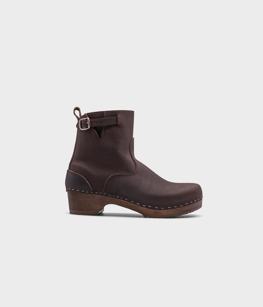 low heeled clog boots in dark brown nubuck leather with a mid-shaft and silver buckle, stapled on a dark wooden base