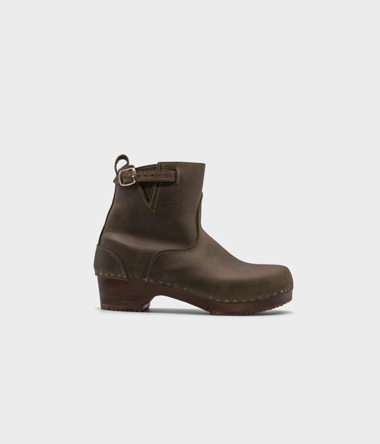 low heeled clog boots in olive green nubuck leather with a mid-shaft and silver buckle, stapled on a dark wooden base