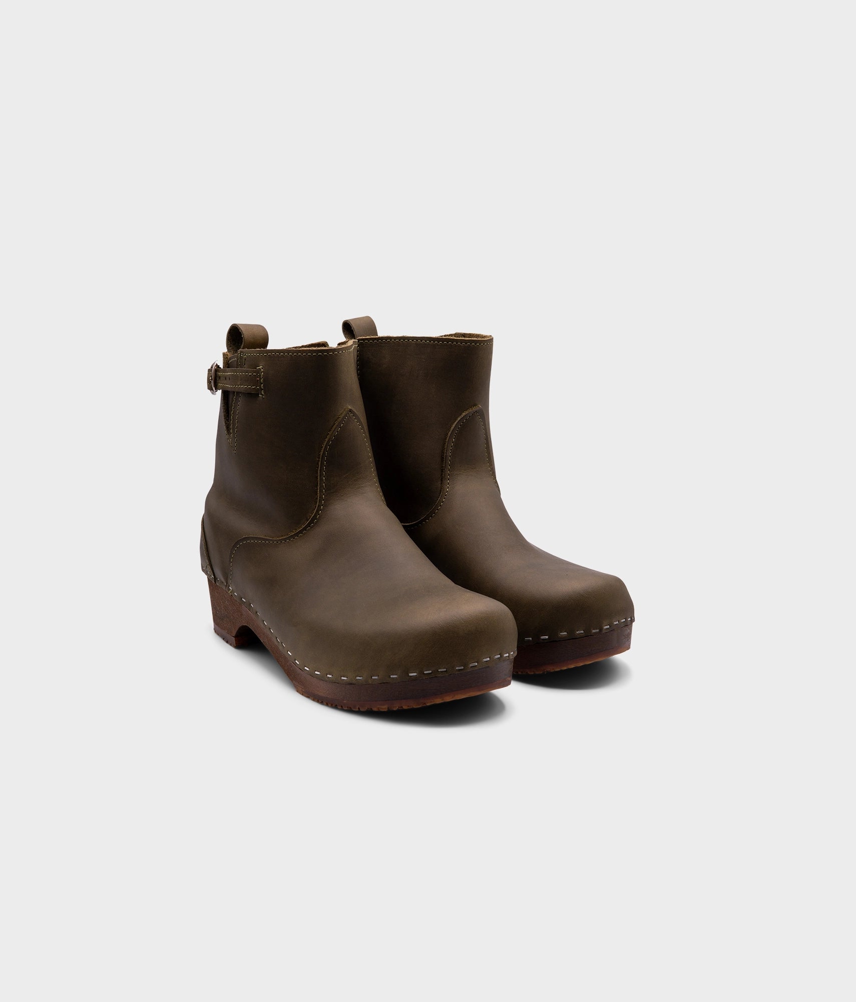 low heeled clog boots in olive green nubuck leather with a mid-shaft and silver buckle, stapled on a dark wooden base