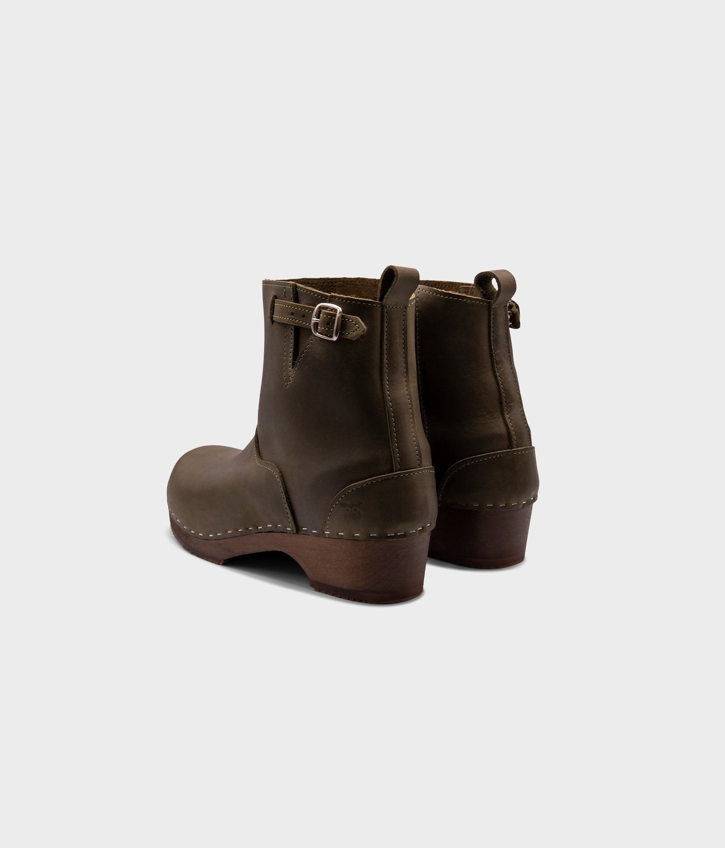 low heeled clog boots in olive green nubuck leather with a mid-shaft and silver buckle, stapled on a dark wooden base