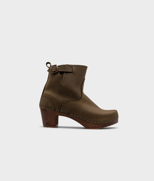 high-heeled clog boots in olive green nubuck leather stapled on a dark wooden base with a silver buckle
