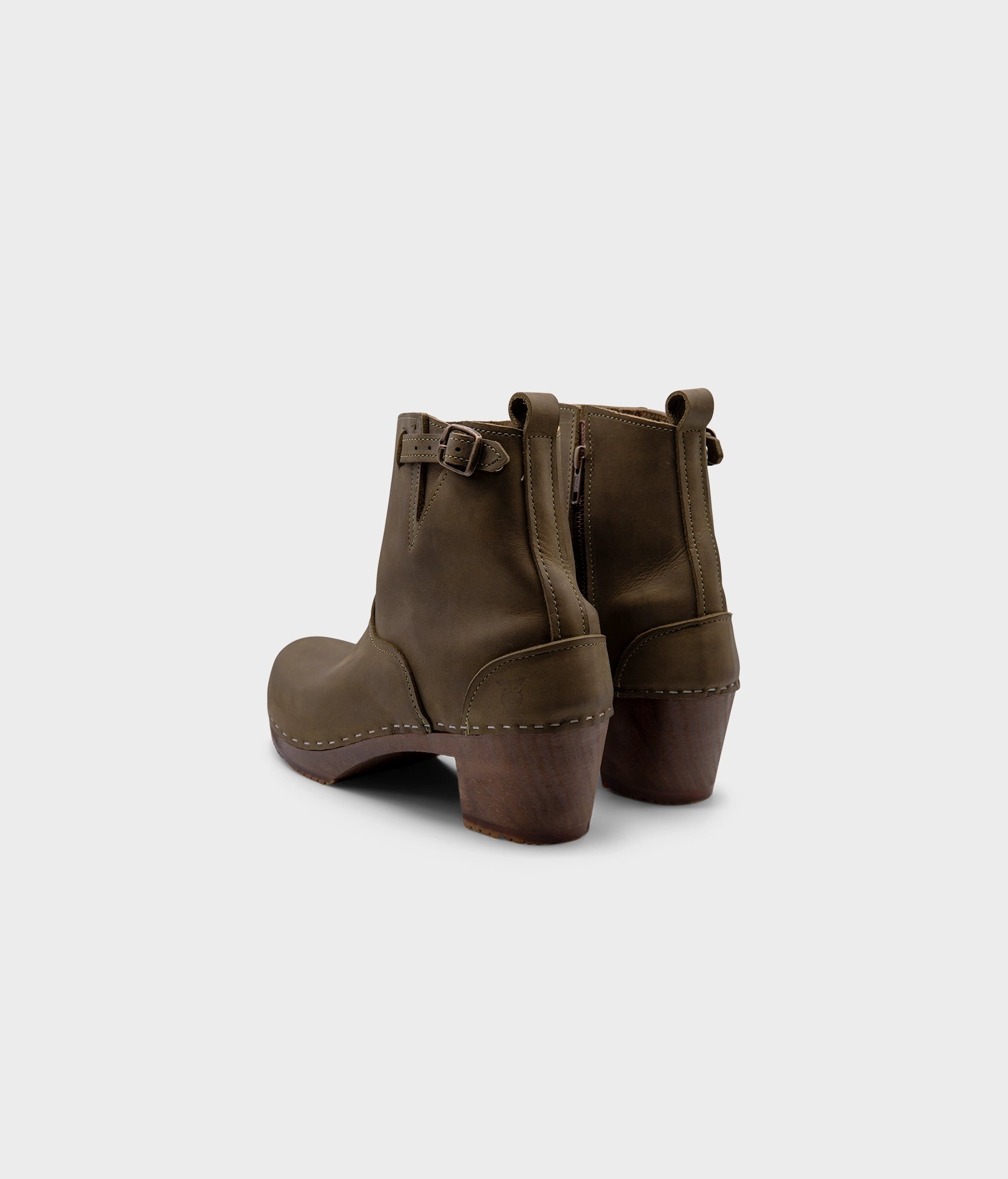 high-heeled clog boots in olive green nubuck leather stapled on a dark wooden base with a silver buckle