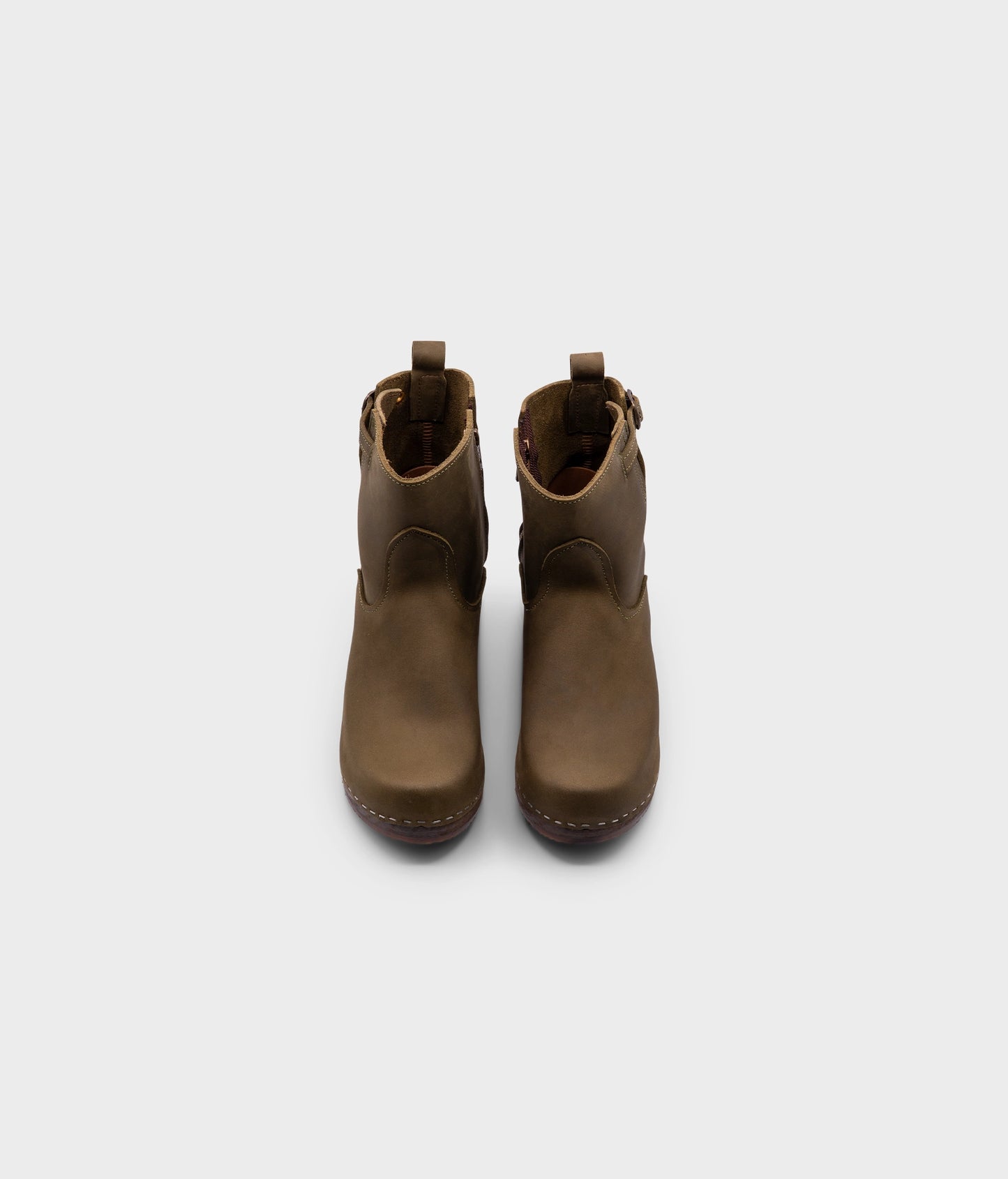 high-heeled clog boots in olive green nubuck leather stapled on a dark wooden base with a silver buckle