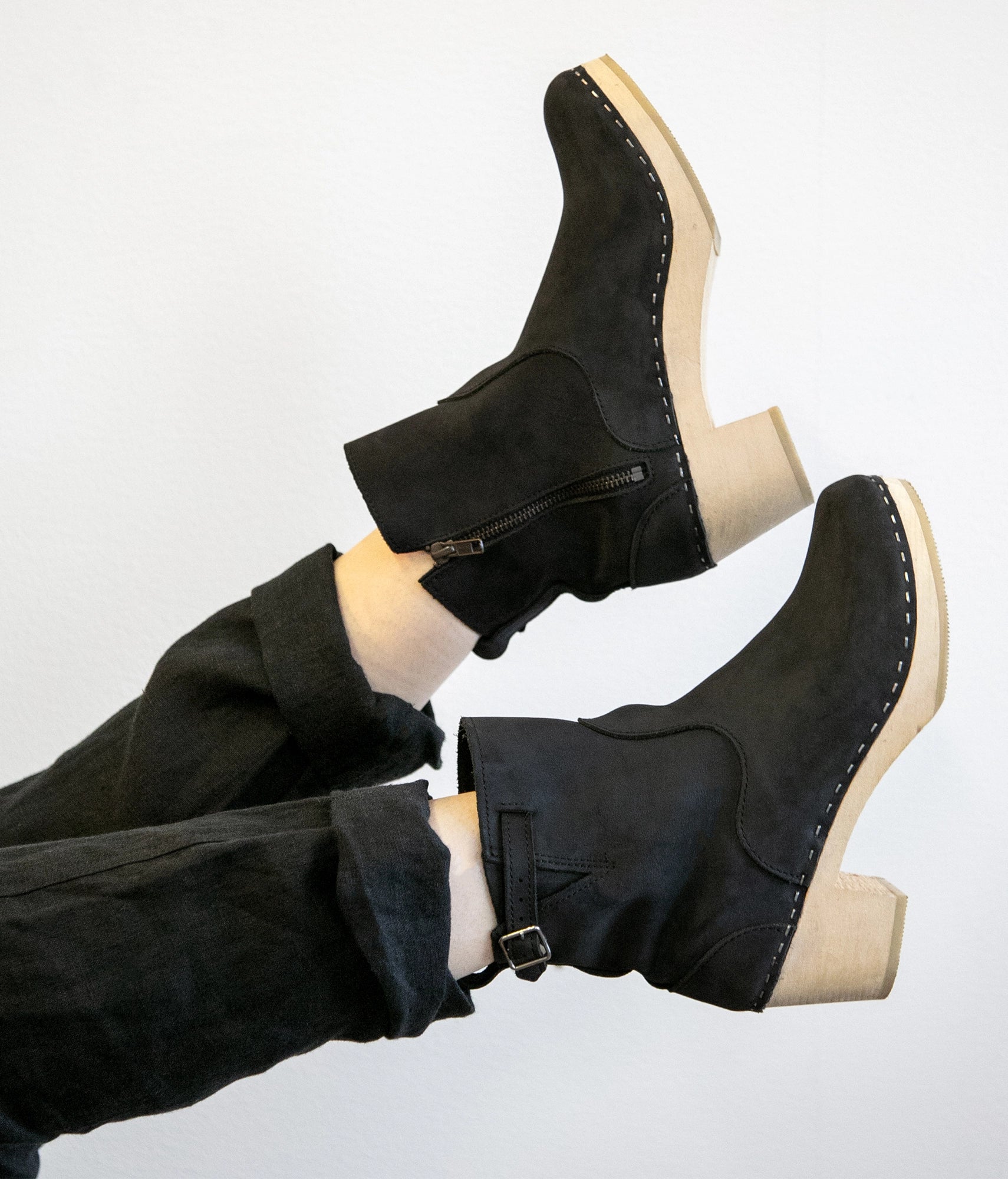 Woman wearing high-heeled clog boots in black nubuck leather stapled on a light wooden base with a silver buckle