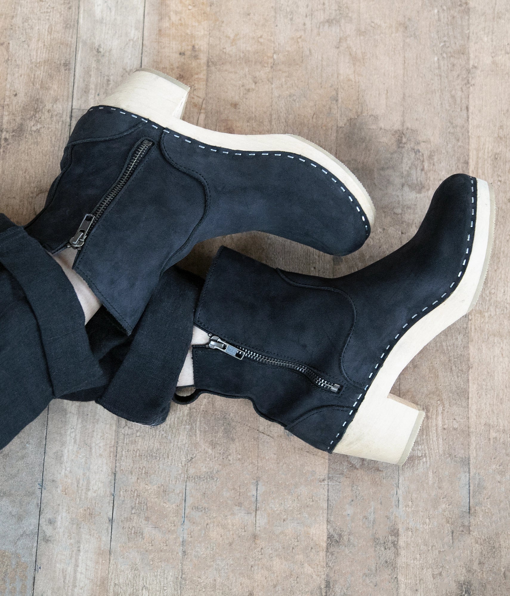 Woman wearing high-heeled clog boots in black nubuck leather stapled on a light wooden base with a silver buckle