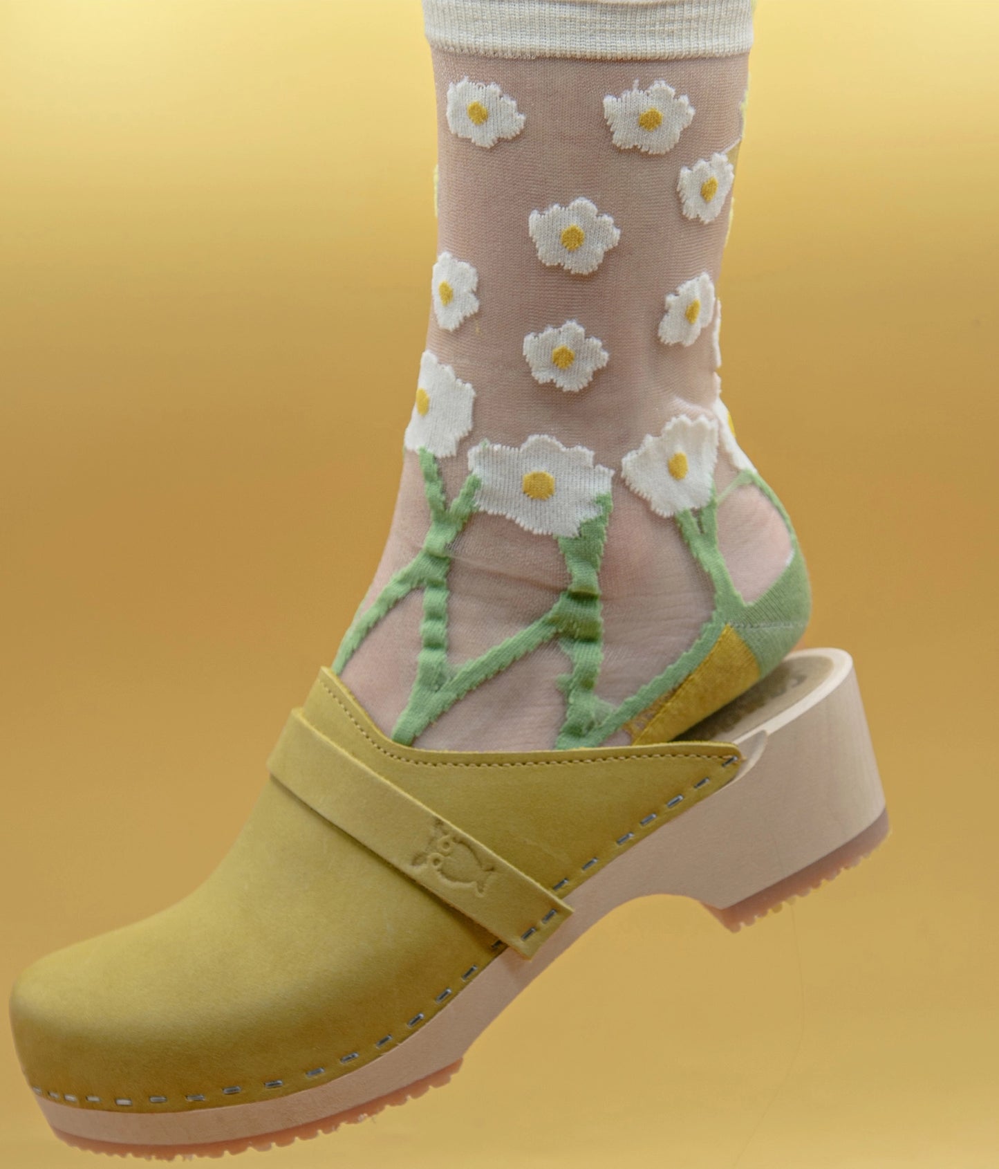 Transparent socks with white flowers and green leaves worn with yellow clogs on a yellow background.classic low heeled clog mule in yellow nubuck leather stapled on a light wooden base with a leather strap