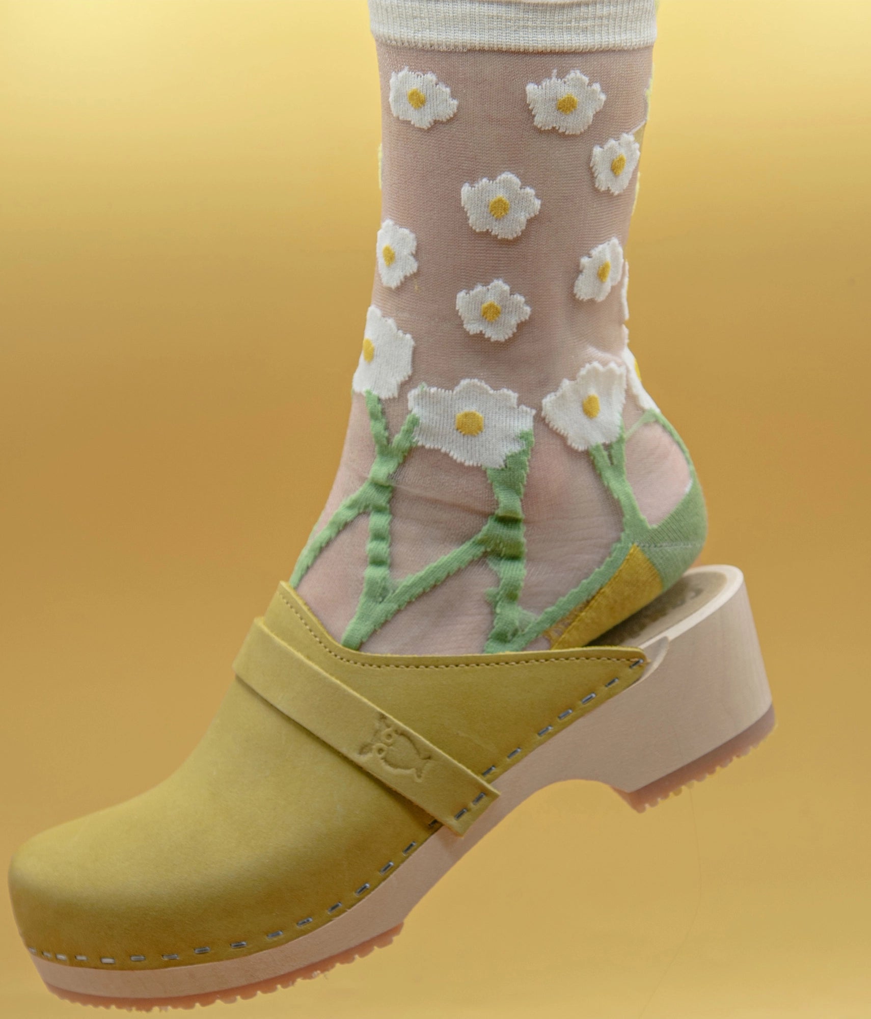Transparent socks with white flowers and green leaves worn with yellow clogs on a yellow background.classic low heeled clog mule in yellow nubuck leather stapled on a light wooden base with a leather strap