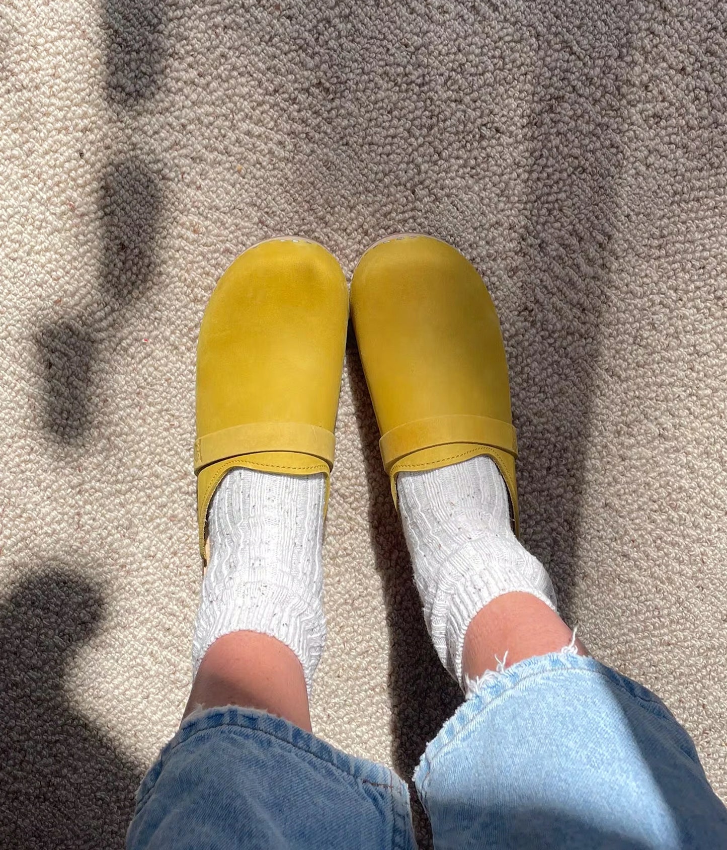 Yellow slippers worn with white socks and blue jeans on a textured surface. classic low heeled clog mule in yellow nubuck leather stapled on a light wooden base with a leather strap