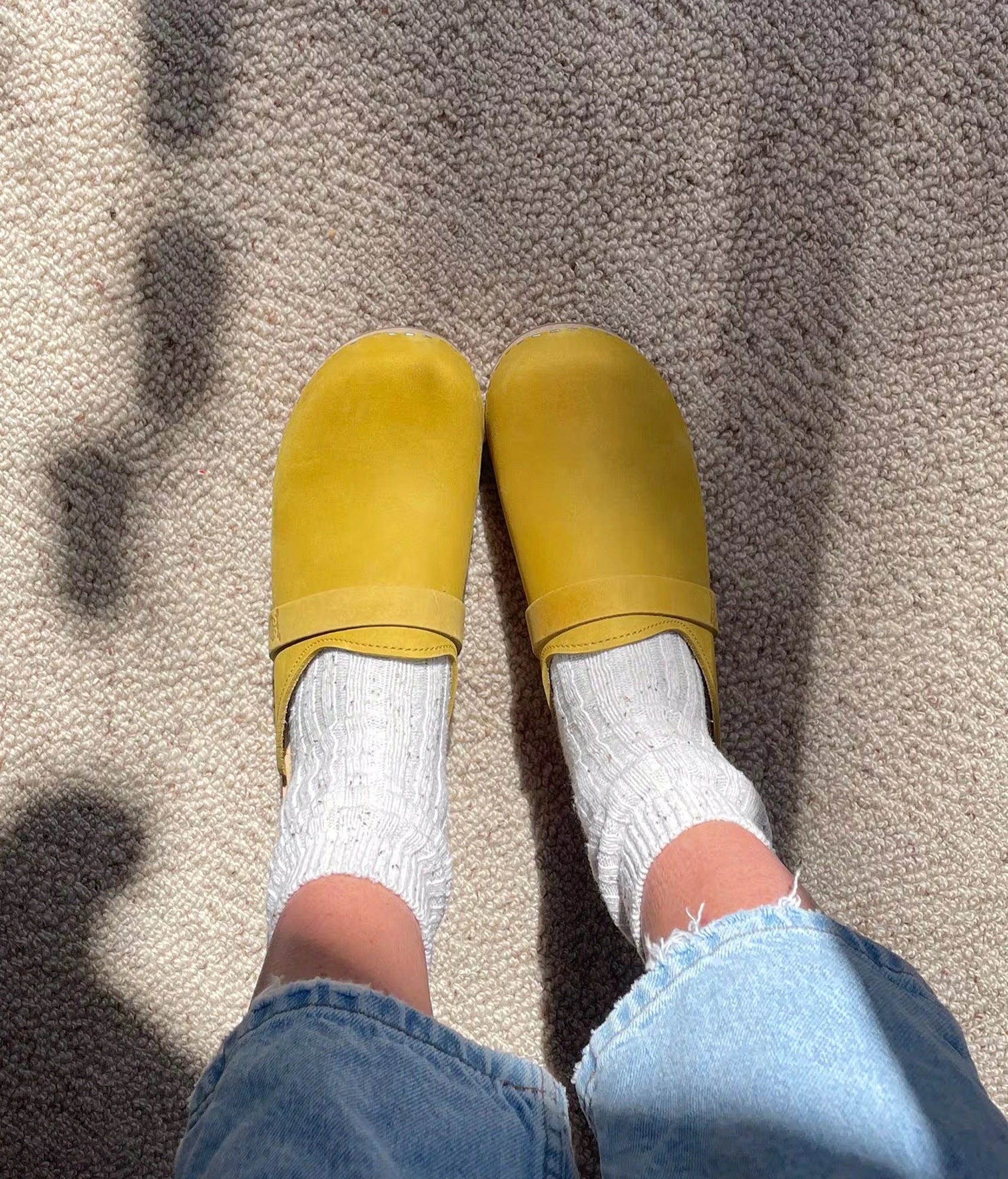 Yellow slippers worn with white socks and blue jeans on a textured surface. classic low heeled clog mule in yellow nubuck leather stapled on a light wooden base with a leather strap
