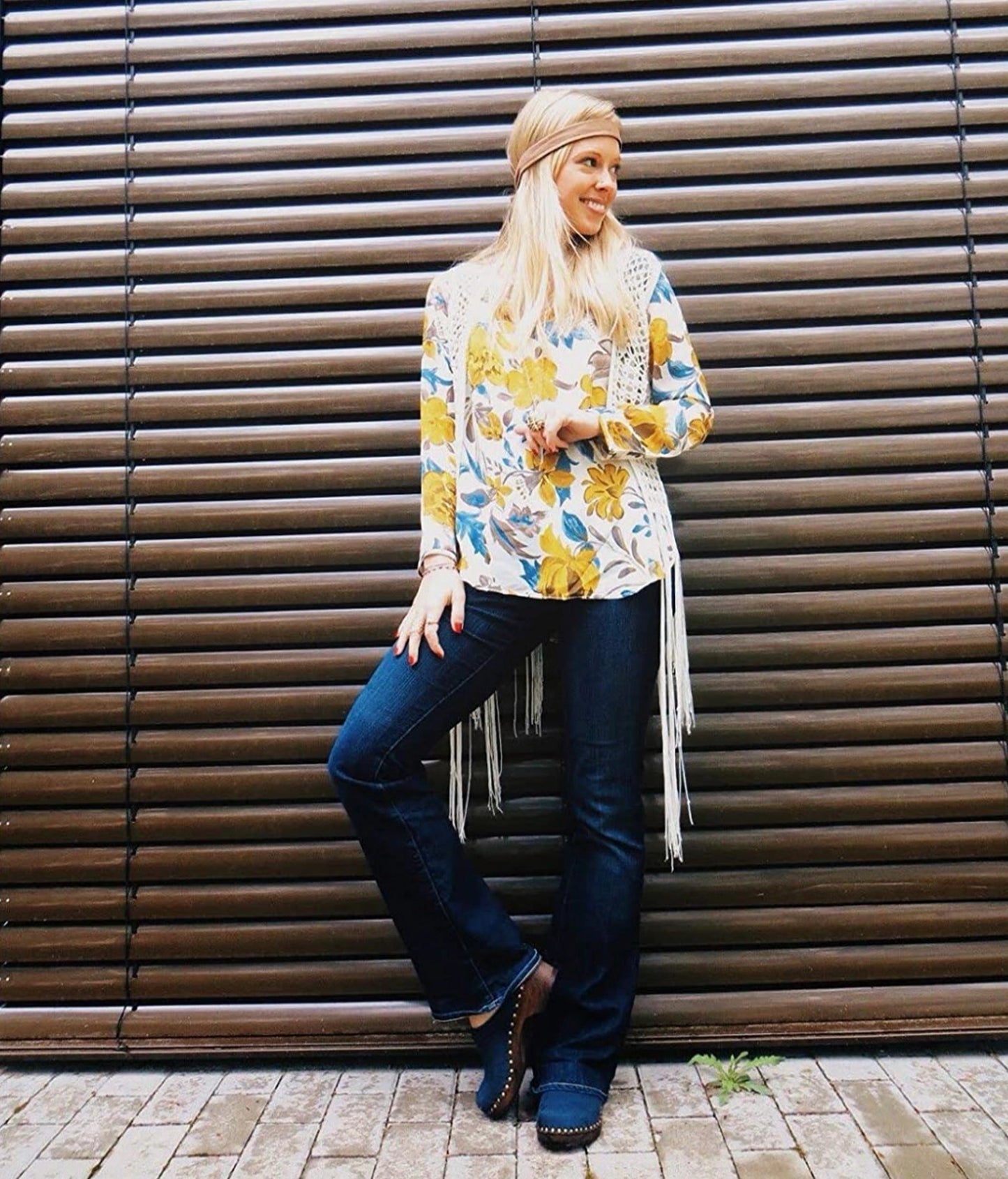 Woman wearing a floral top and blue jeans leaning against a wooden wall. low heeled clog mules in navy nubuck leather stapled on a dark wooden base with brass gold studs