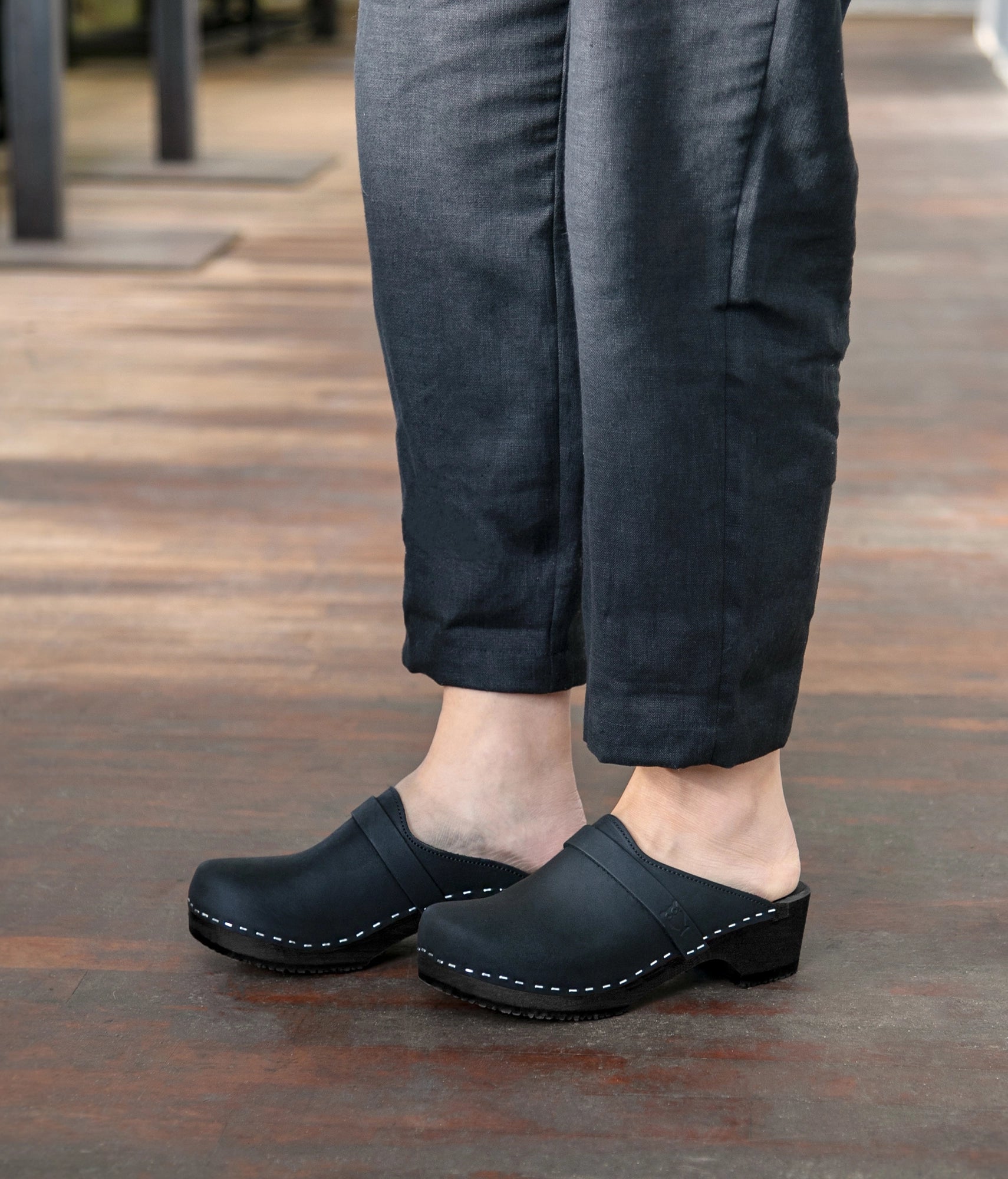 Person wearing black clogs and dark pants on a wooden floor. Classic low heeled clog mule in black nubuck leather stapled on a black wooden base with a leather strap