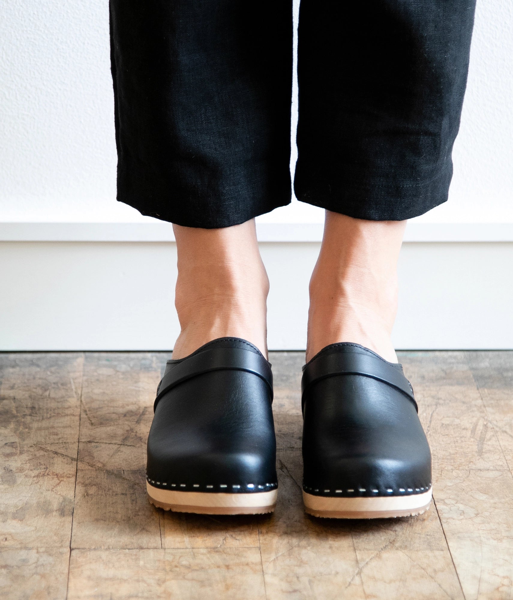 Person wearing black leather clogs on a wooden floor.