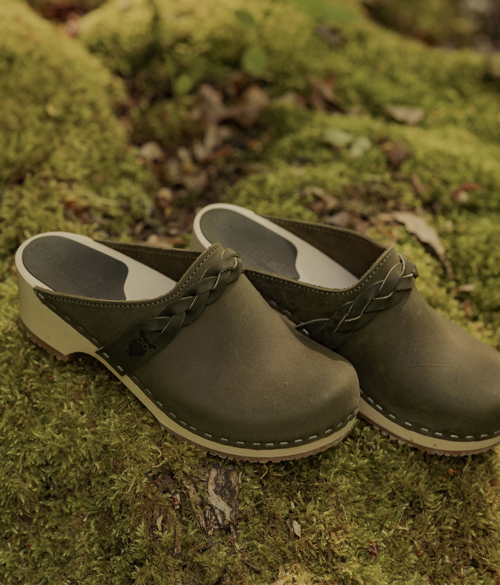 low heeled clog mules in olive green nubuck leather with a braided strap stapled on a light wooden base on mossy surface
