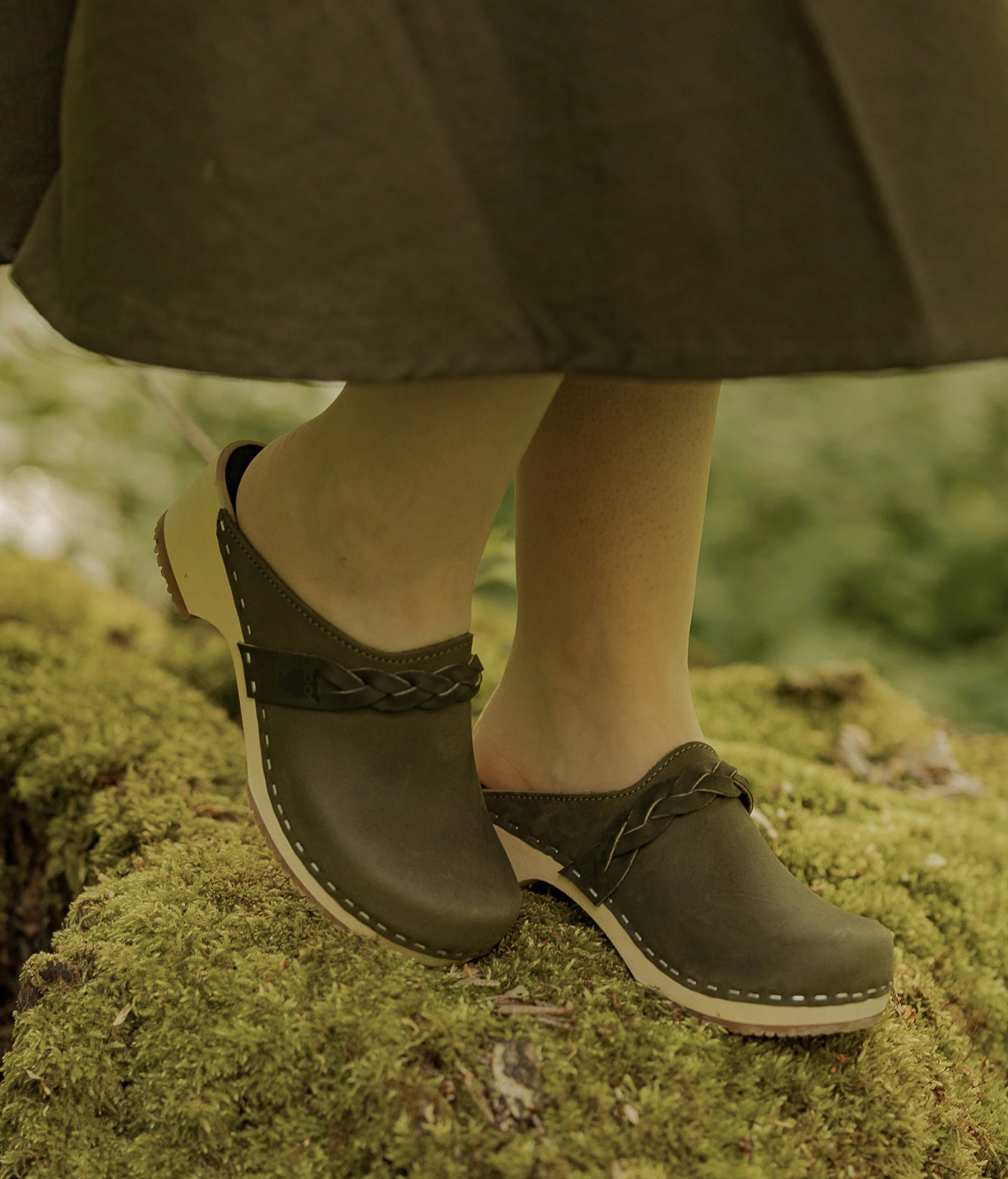 Person wearing green clogs on a mossy surface. Low heeled clog mules in olive green nubuck leather with a braided strap stapled on a light wooden base 