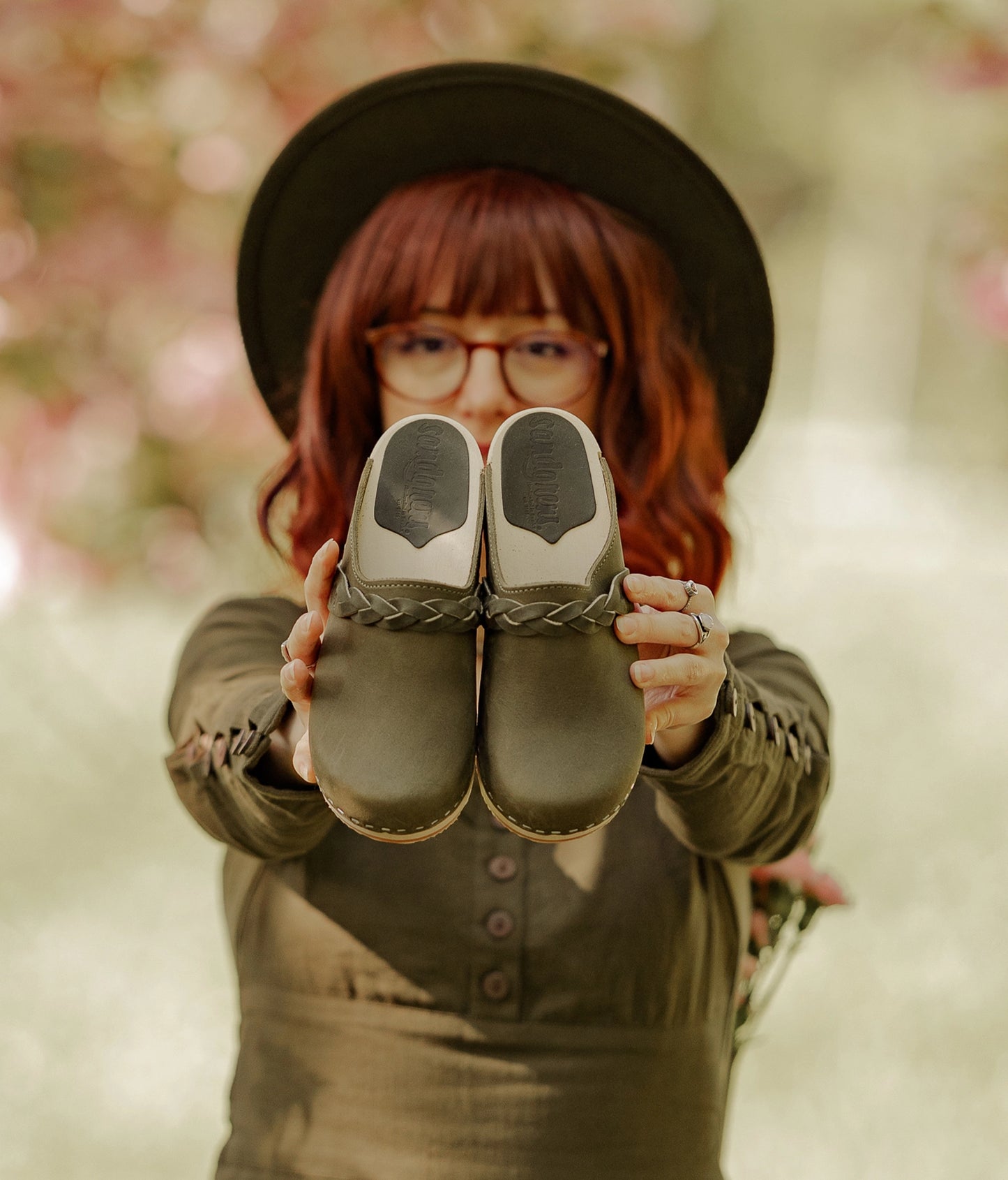 Person holding a pair of shoes in front of a blurred natural background. Low heeled clog mules in olive green nubuck leather with a braided strap stapled on a light wooden base